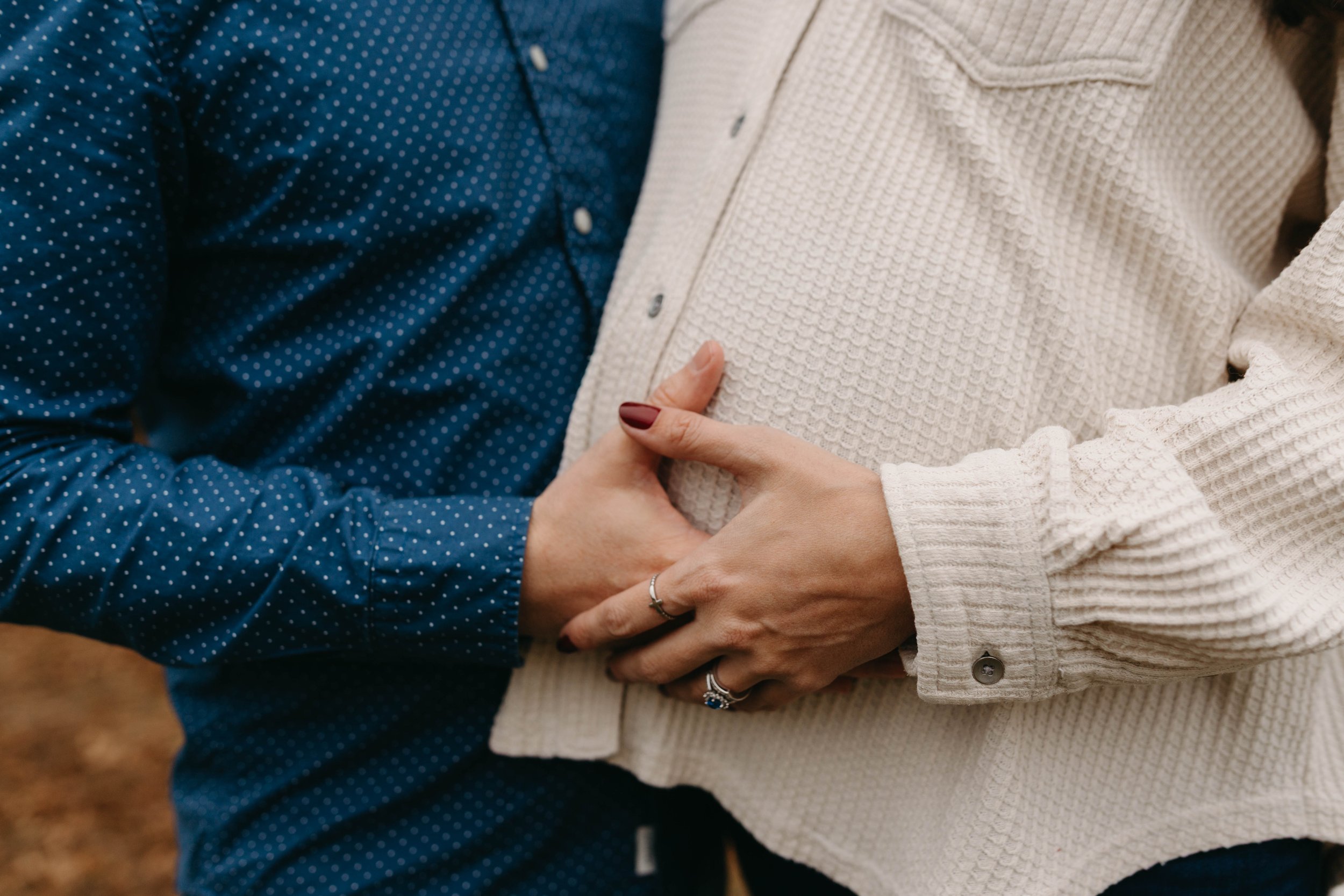 Two people standing close, holding hands over a pregnant belly, one person wearing a blue polka dot shirt and the other in a cream textured sweater.