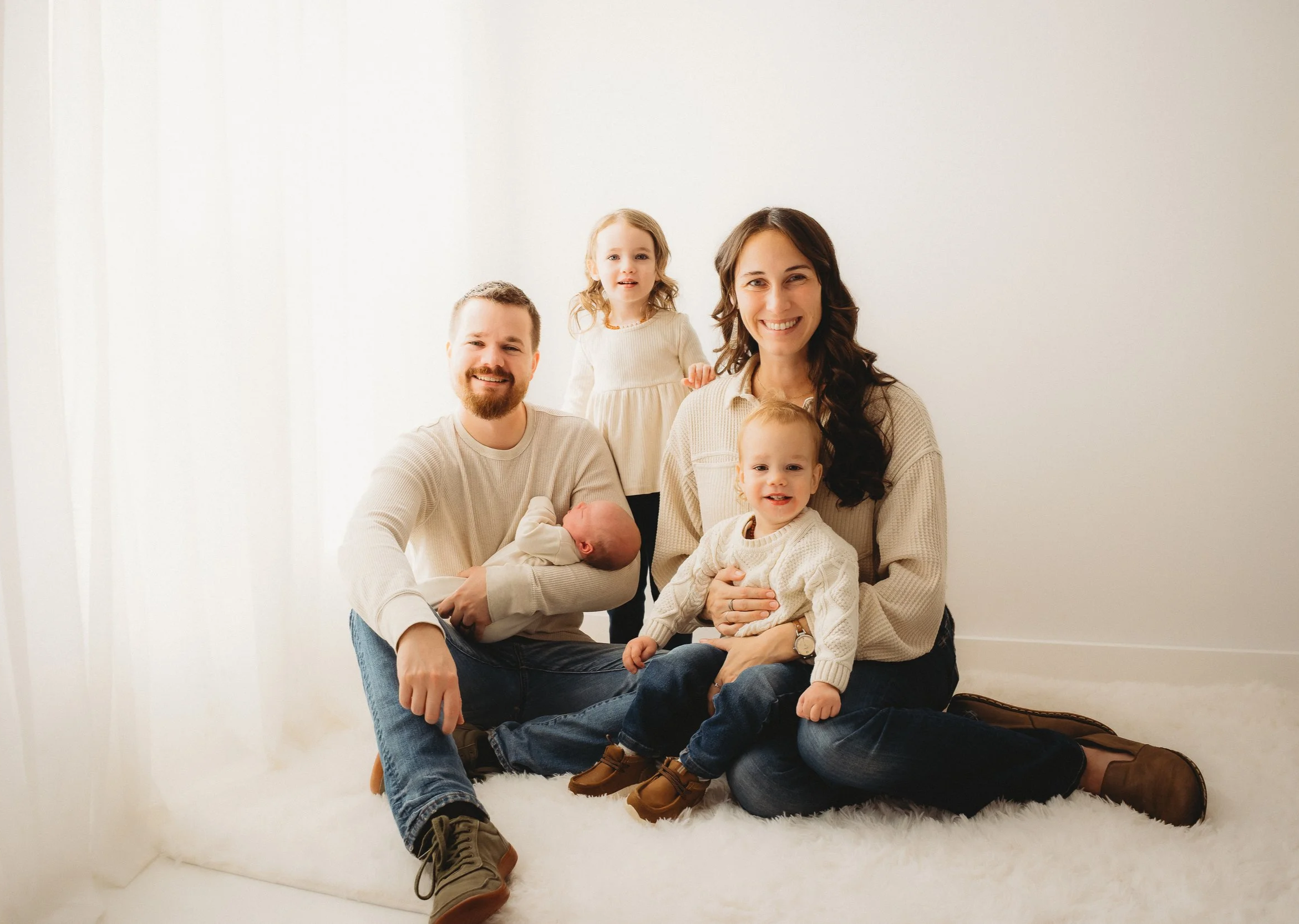 Family portrait of a man, woman, and three children sitting together on a white fuzzy rug in a bright room with white curtains. The man is holding a newborn, the woman is holding a toddler, and the other child stands behind them, all smiling.