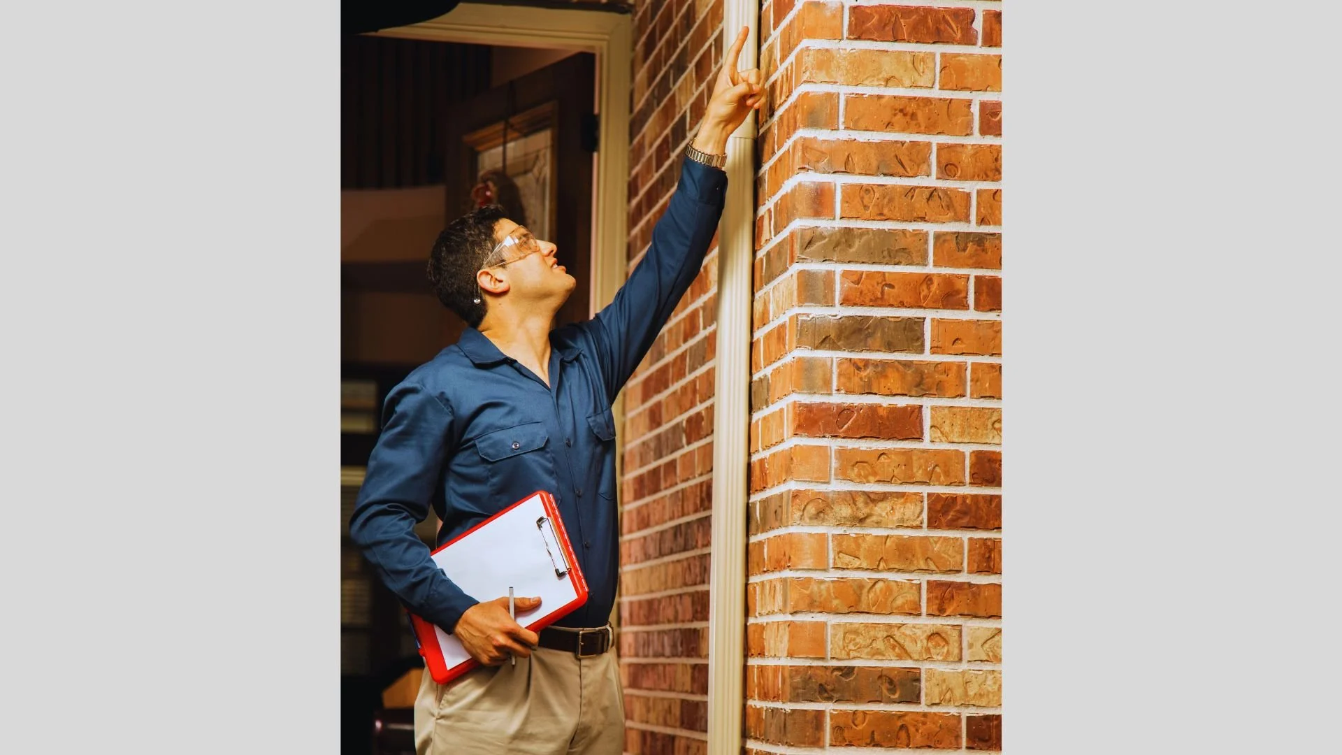 Une femme en blouse bleue tient un clipboard rouge et un stylo, et pointe vers un mur en briques, probablement en train de faire une inspection ou une évaluation.