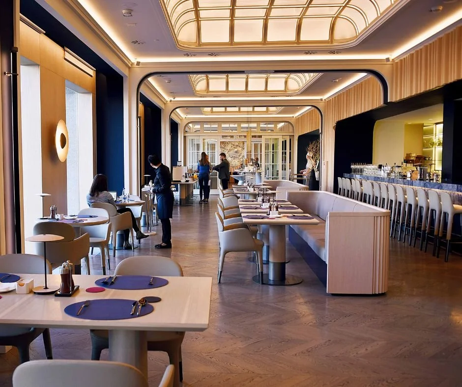 An elegant restaurant with beautifully set tables, modern chairs, and waiters taking orders. Large windows let in plenty of natural light, creating a warm and sophisticated atmosphere.