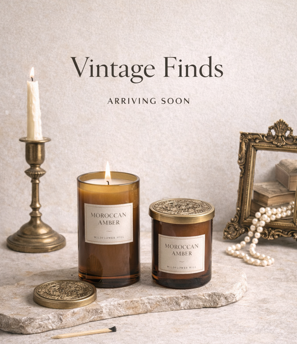 Photo of two Moroccan Amber scented candles, one lit, on a stone slab, with vintage decor including a candlestick, framed picture, and pearl necklace, and a message reading 'Vintage Finds ARRIVING SOON' in the background.