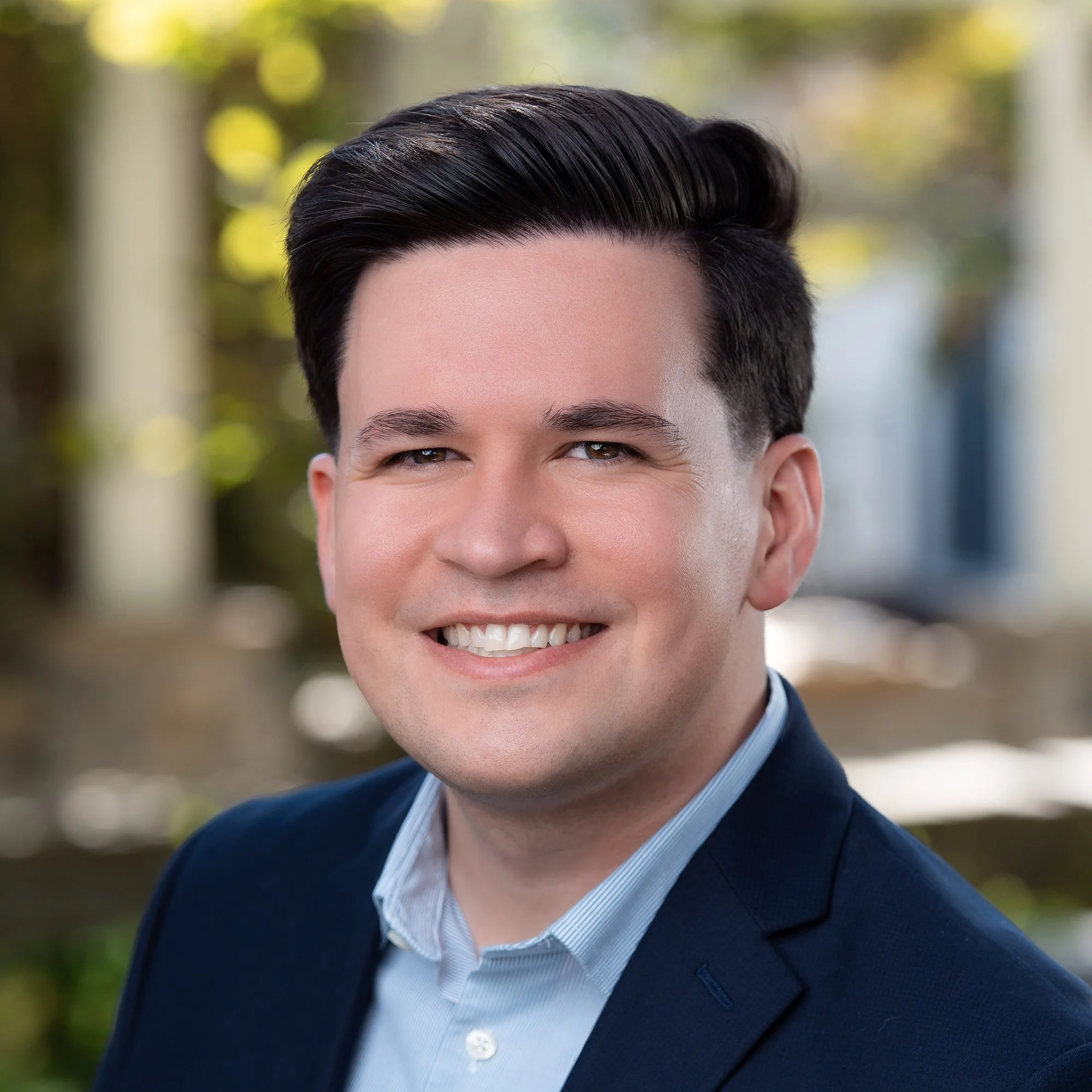 Nick Pomes smiling in a navy blazer and light blue shirt poses outdoors with blurred greenery in the background.