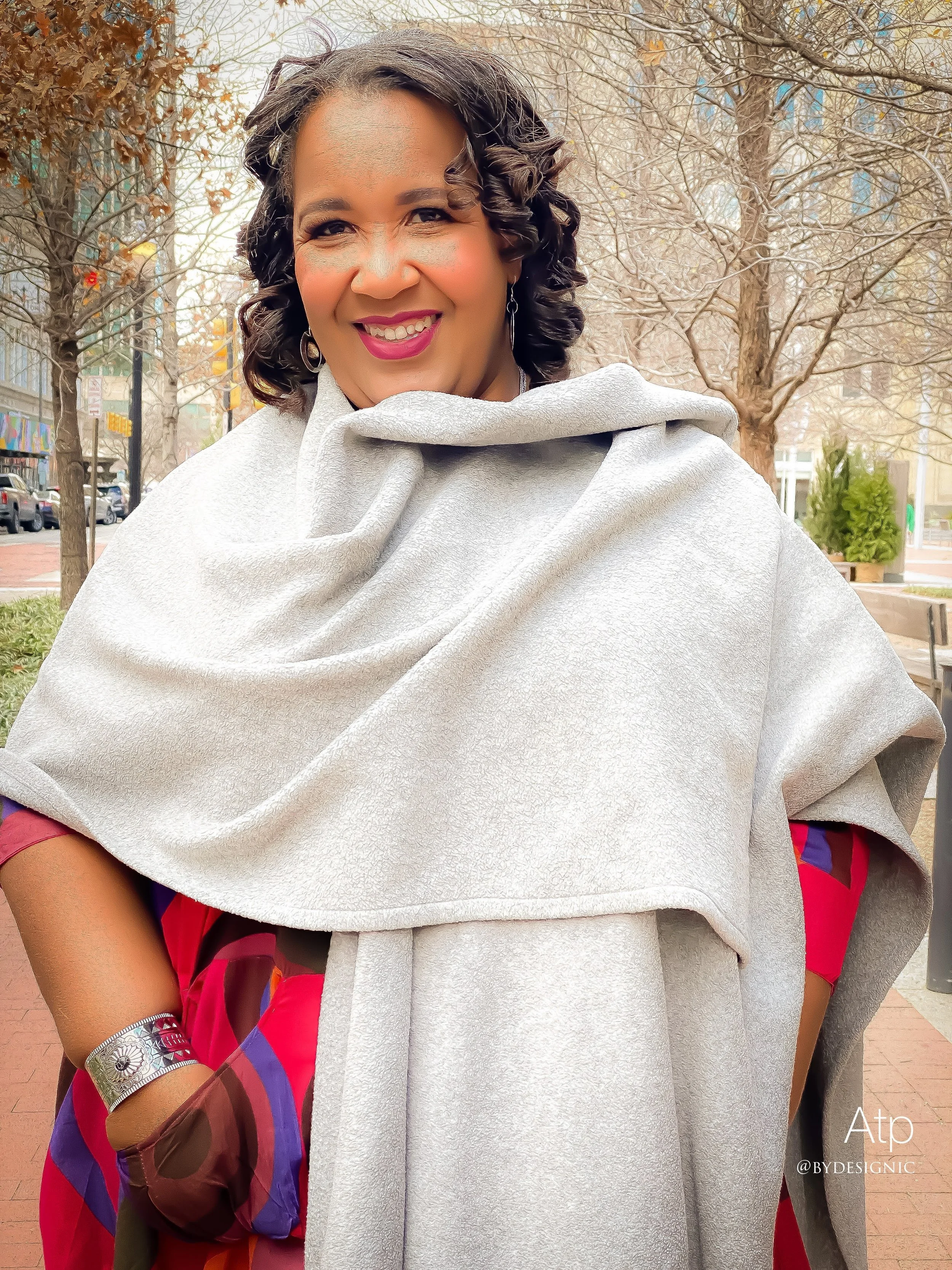 A woman with curly dark hair smiling, wearing a gray shawl and a red and purple dress, standing outdoors in a city park with leafless trees and buildings in the background.