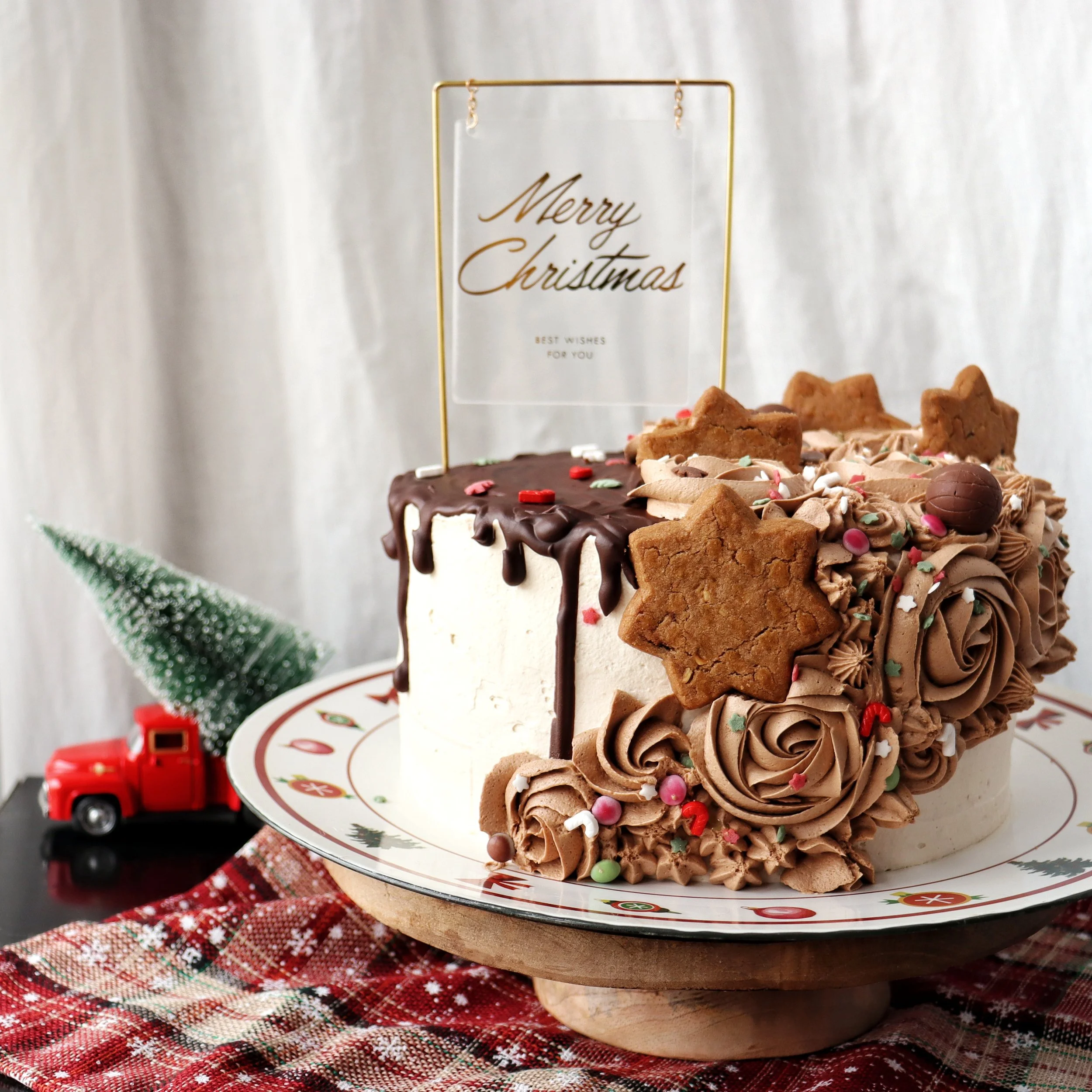 A Christmas cake decorated with chocolate, cookies, sprinkles, and a 'Merry Christmas' topper. A small green Christmas tree and a red toy truck are in the background. The cake is on a festive plate on top of a red plaid tablecloth.