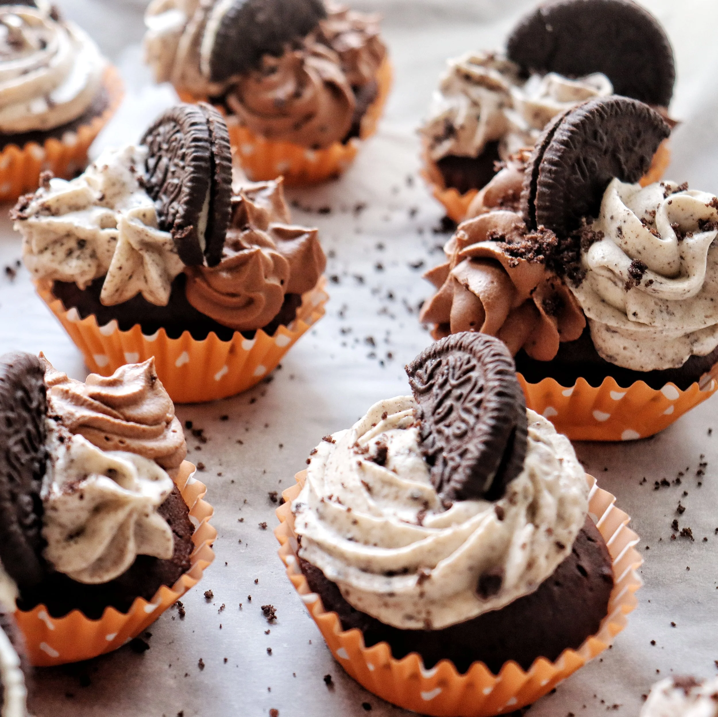Chocolate cupcakes with cream cheese and whipped cream frosting topped with Oreo cookies, arranged on a sheet with crumbs scattered around.