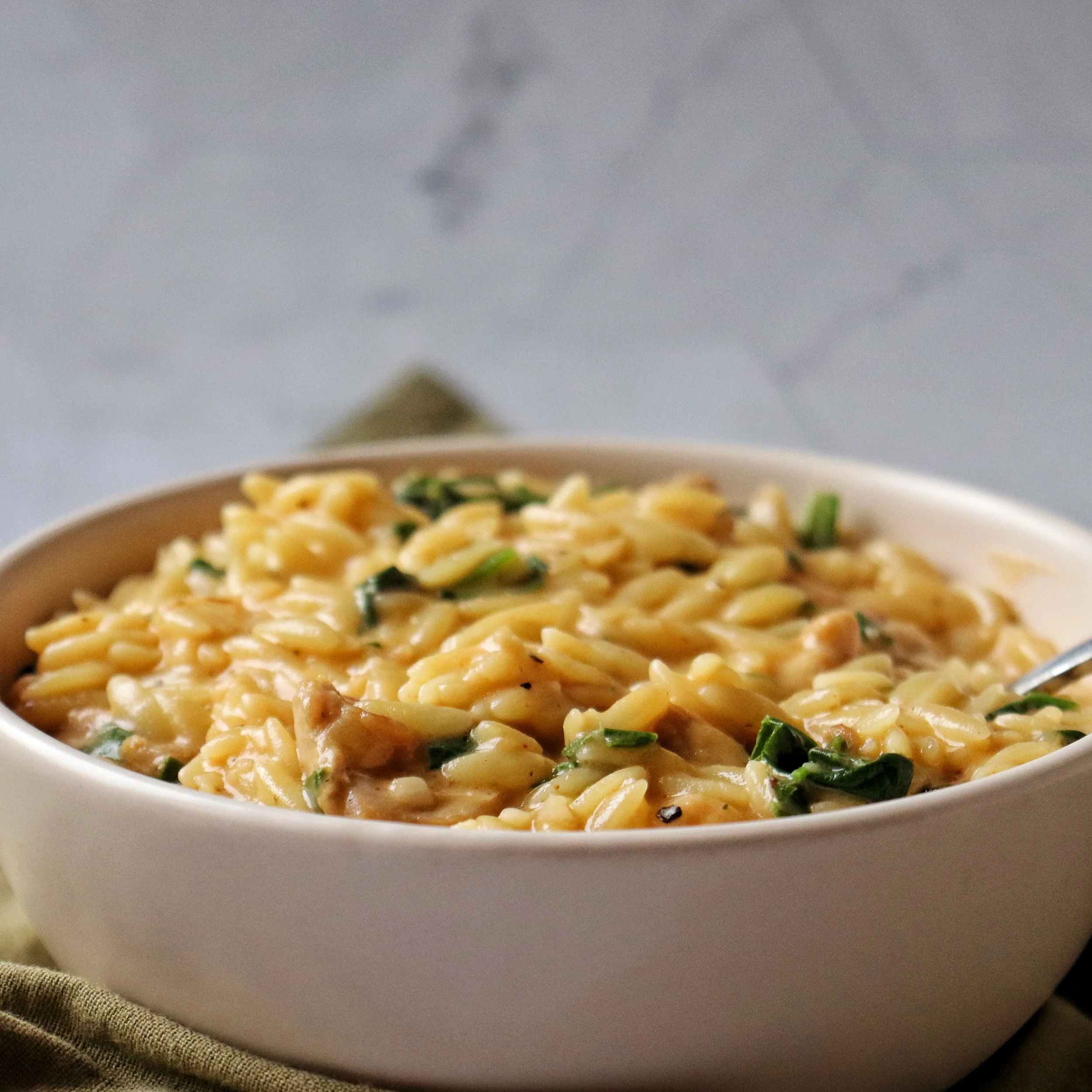 A white bowl filled with creamy chicken and orzo pasta, placed on a beige cloth with a fork partially submerged in the dish.
