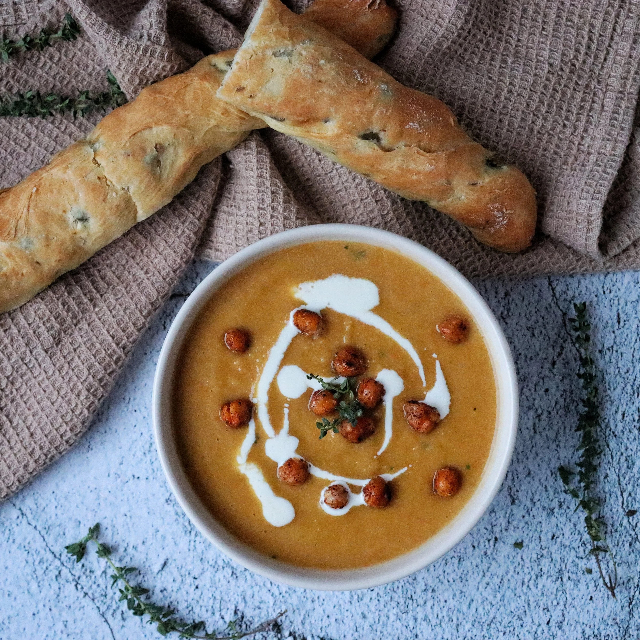 A bowl of creamy pumpkin soup garnished with a drizzle of cream, roasted chickpeas, and a sprig of thyme, accompanied by two slices of olive bread.