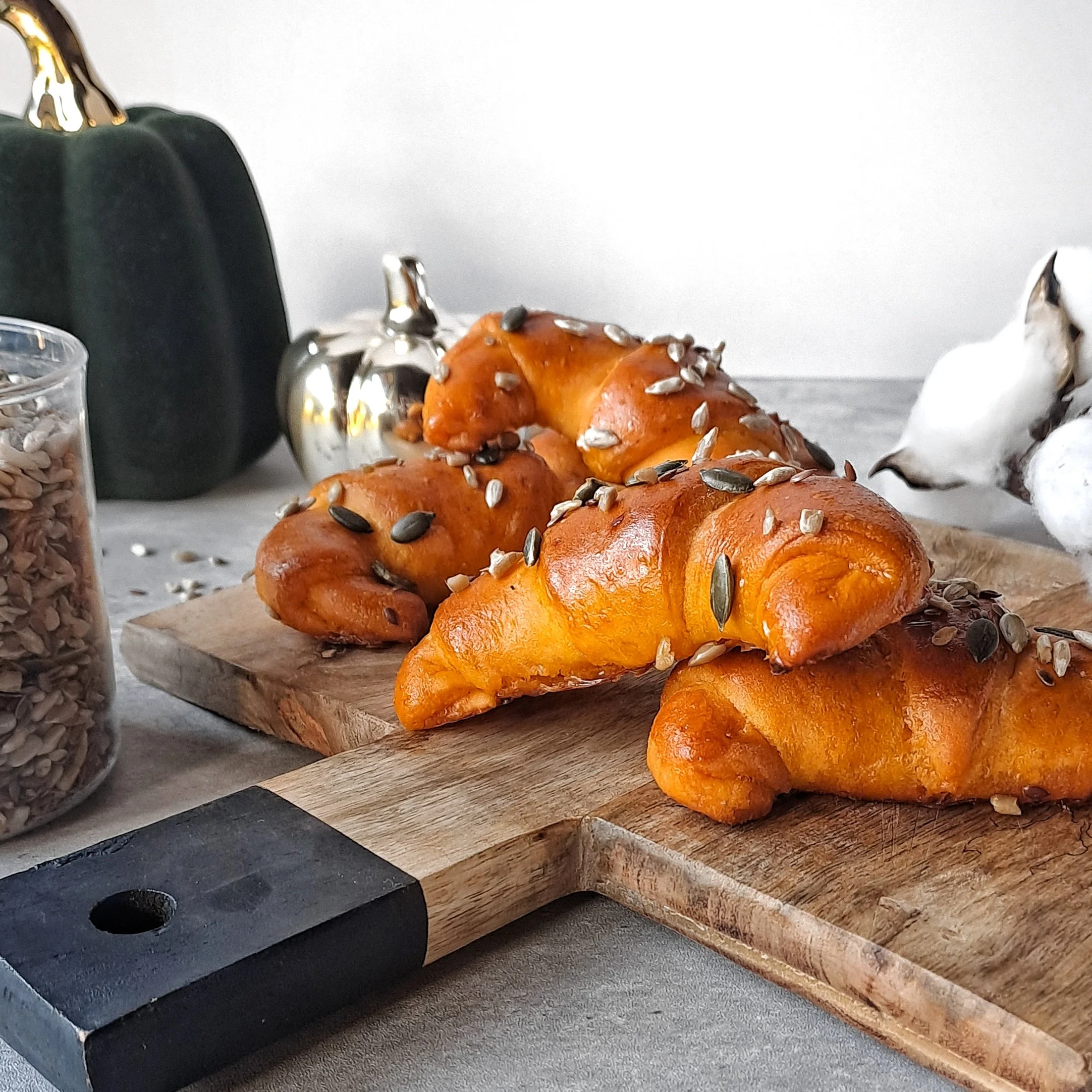 Three golden-brown pumpkin croissants topped with sunflower seeds on a wooden cutting board.