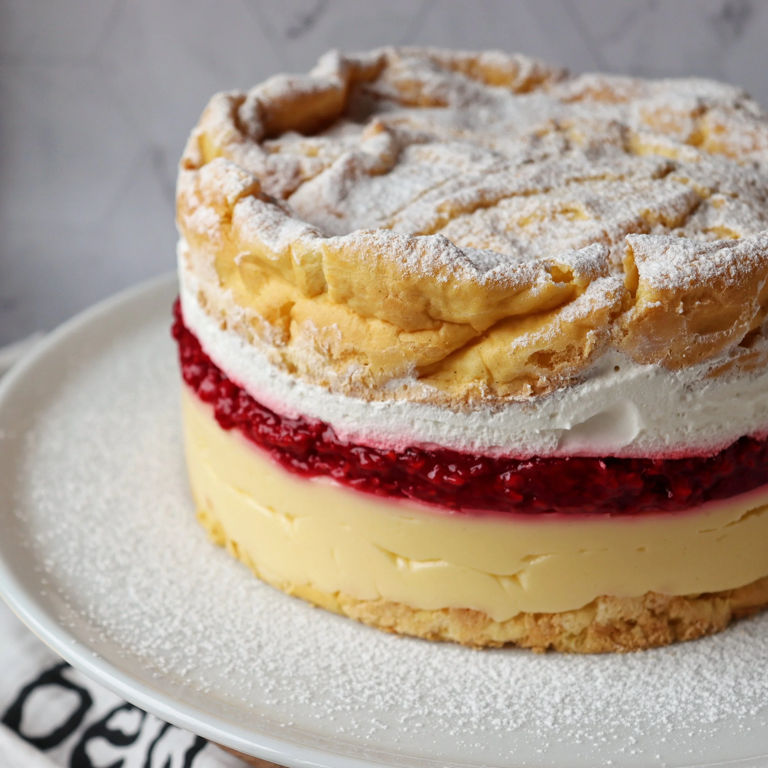 A layered Karpathian Mountain cake dessert with a crumbly top dusted with powdered sugar, a layer of red fruit filling, and a layer of creamy yellow custard, served on a white plate.