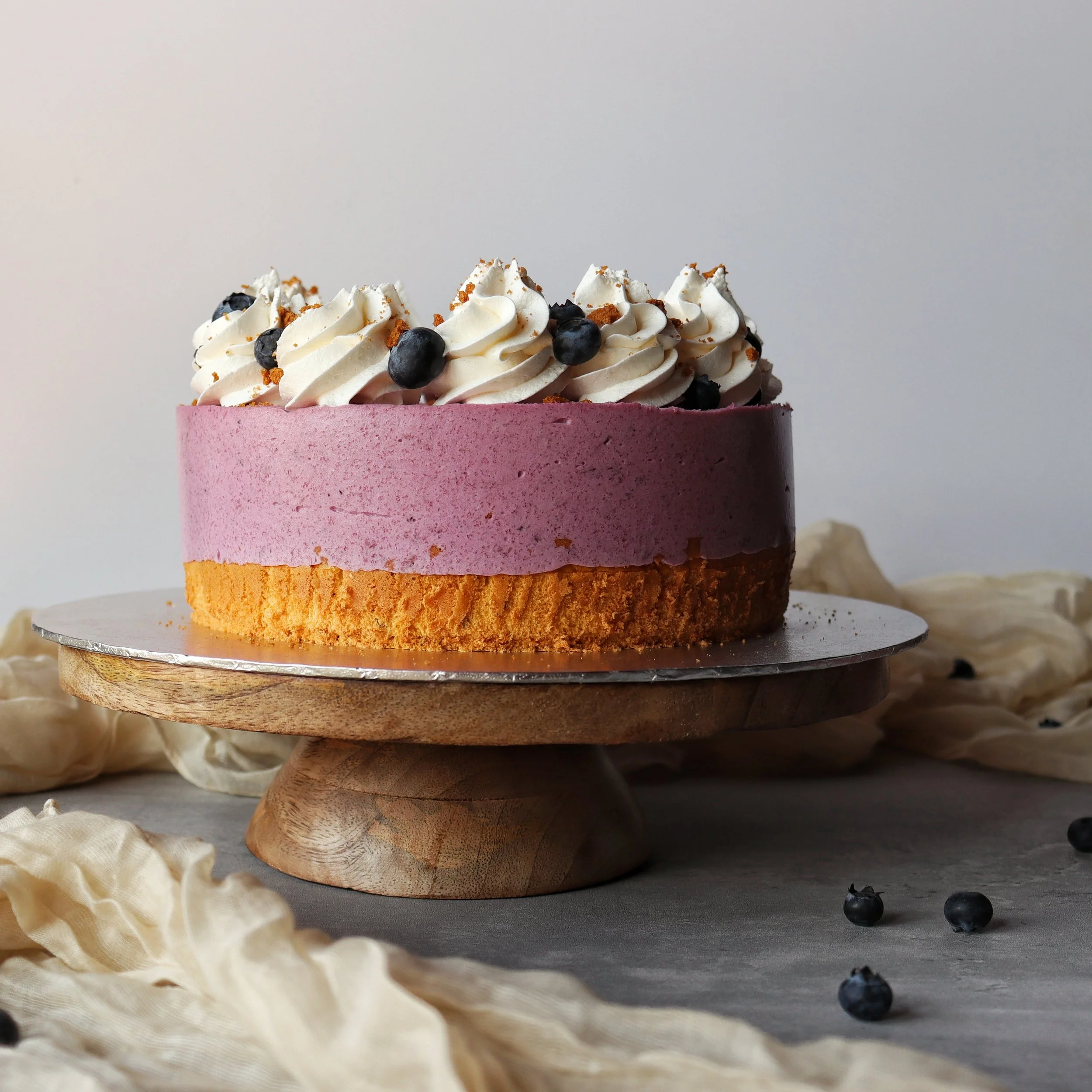 A layered berry mousse cake with whipped cream and blueberries on top, sitting on a wooden cake stand, surrounded by scattered blueberries and light-colored fabric.