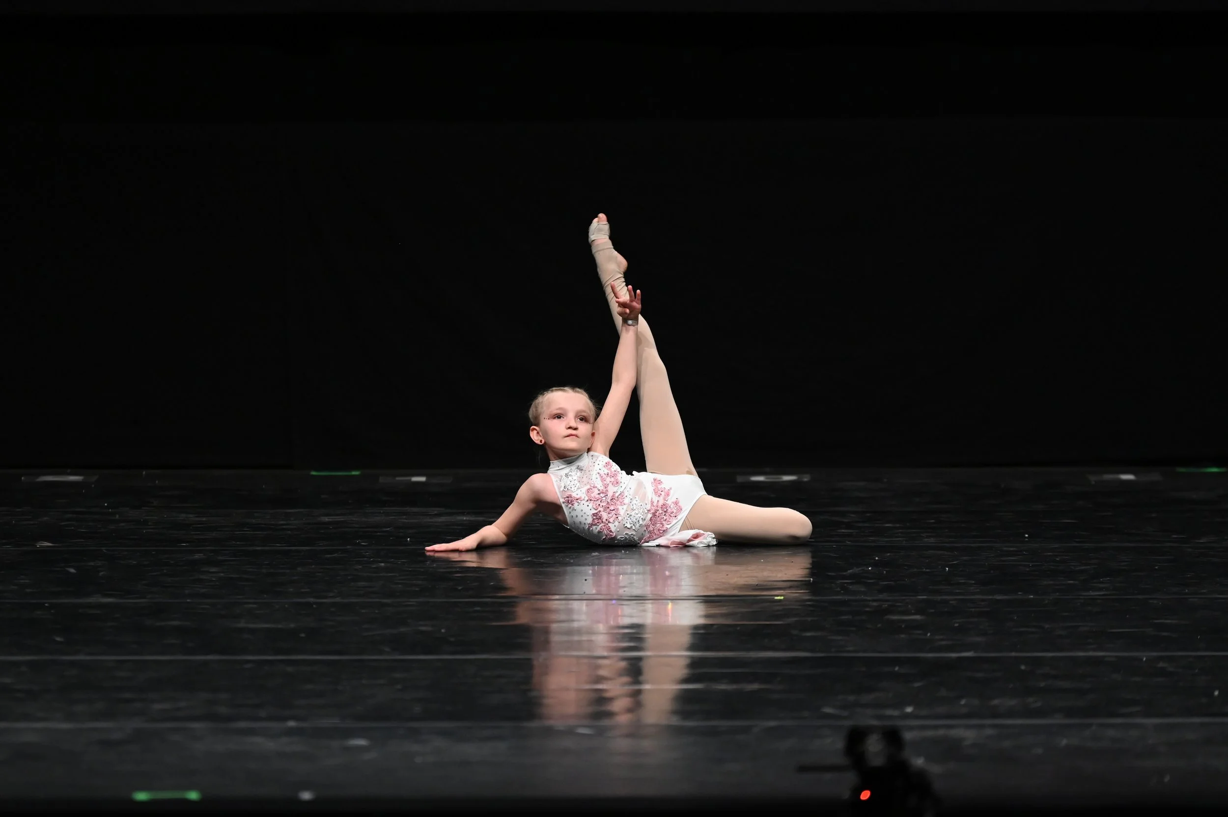 Ein junges Mädchen beim Ballett auf der Bühne, liegt auf dem Boden, hebt ein Bein hoch und hält es mit einer Hand, während sie die andere Hand flach auf die Bühne legt.