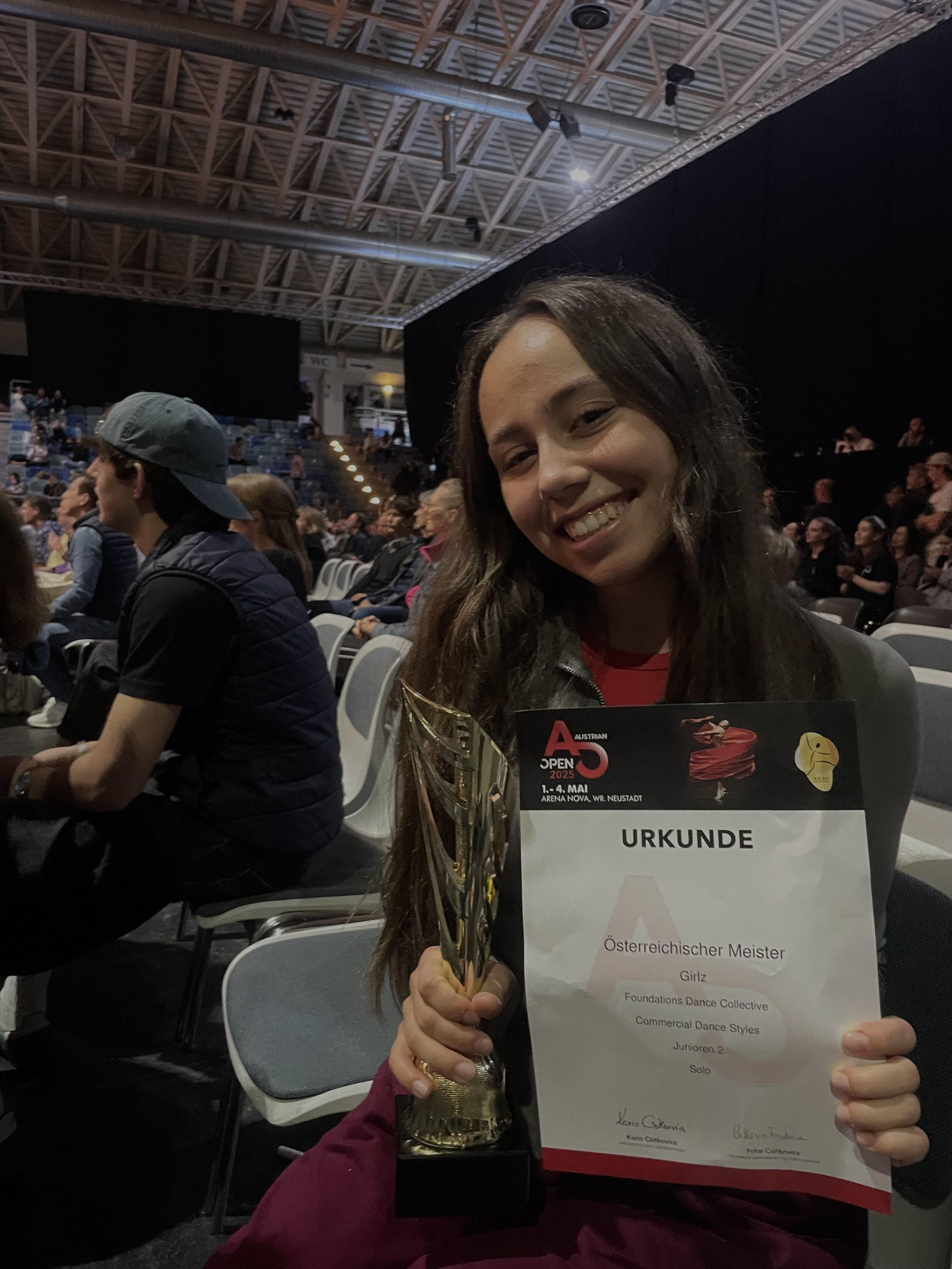 Chiara Adler steht alleine bei den Austrian Open und hält ihre Trophäe sowie ihr Zertifikat in den Händen. Ihr Ausdruck zeigt Stolz und Zufriedenheit nach ihrem erfolgreichen Auftritt.