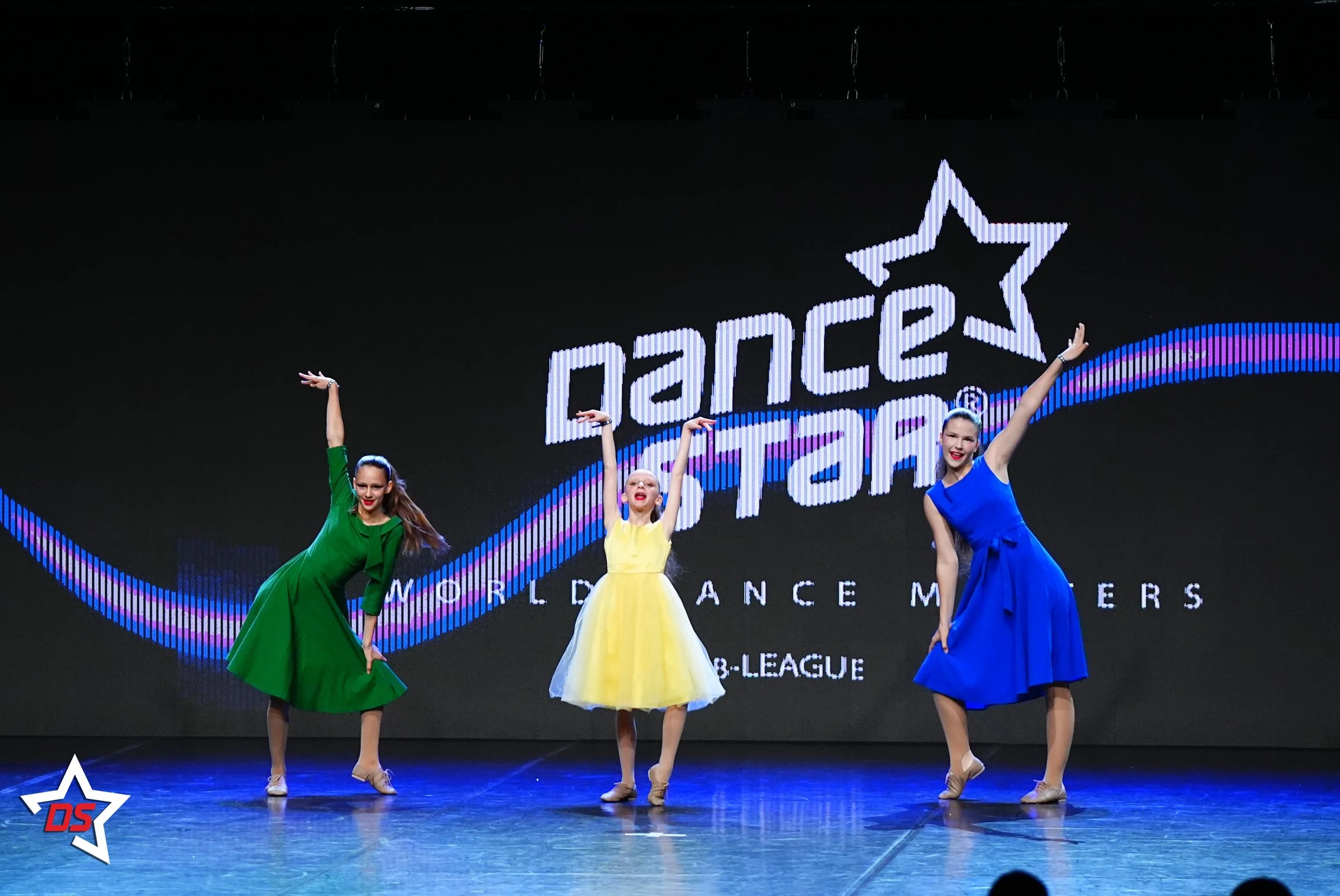 Tänzerinnen in farbigen Kleidern (grün, gelb, blau) auf einer Bühne bei einer Tanzperformance, mit einem großen LED-Bildschirm im Hintergrund, der das Disc-Logo und den Text "World Dance Masters" und "B-League" zeigt.