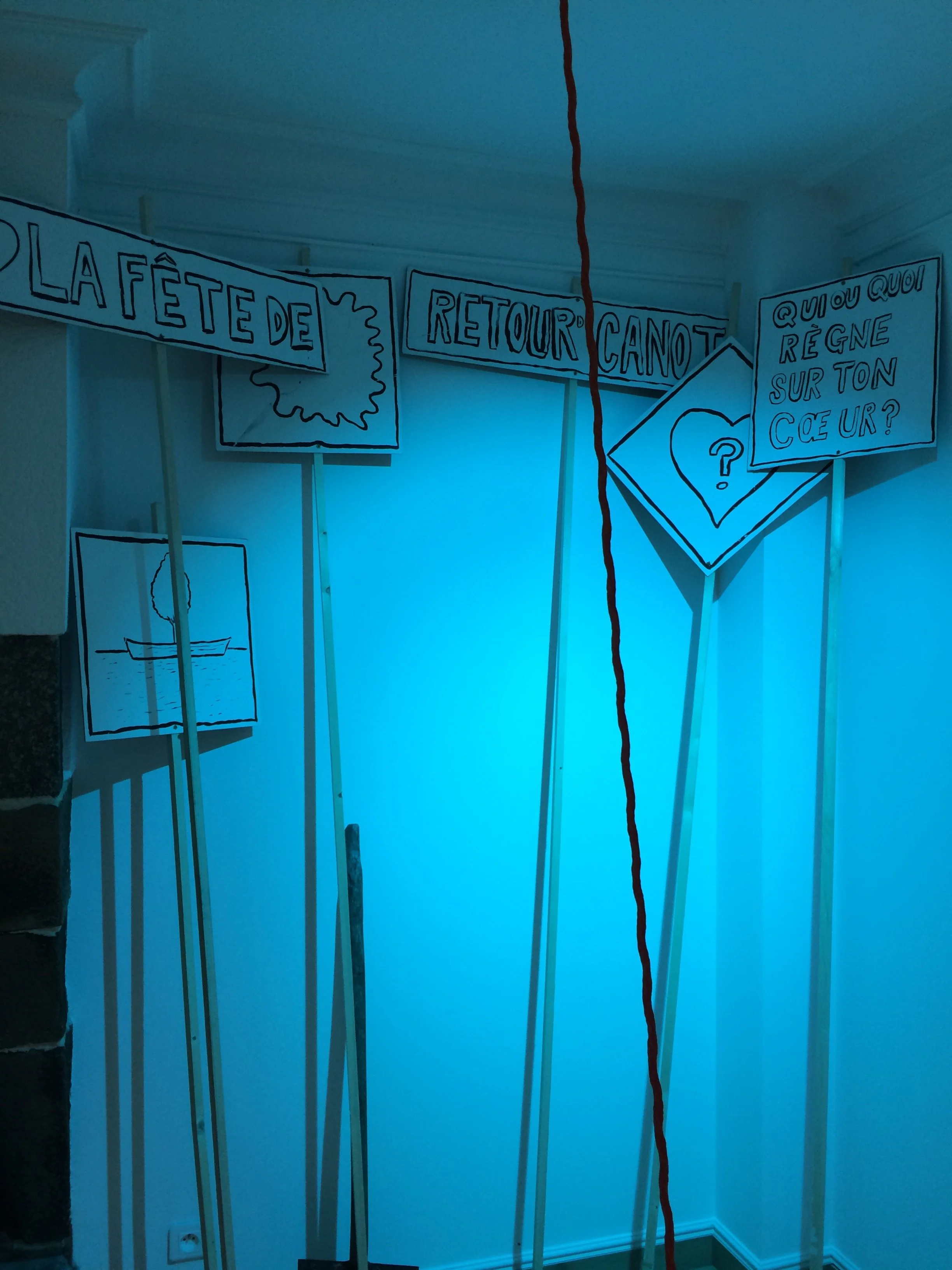 Décorations en papier avec des textes en français, incluant 'La fête de', 'retour canot', et 'qu'est ce qui régne sur ton cœur ?', fixées sur des bâtons dans une pièce avec un éclairage bleu.