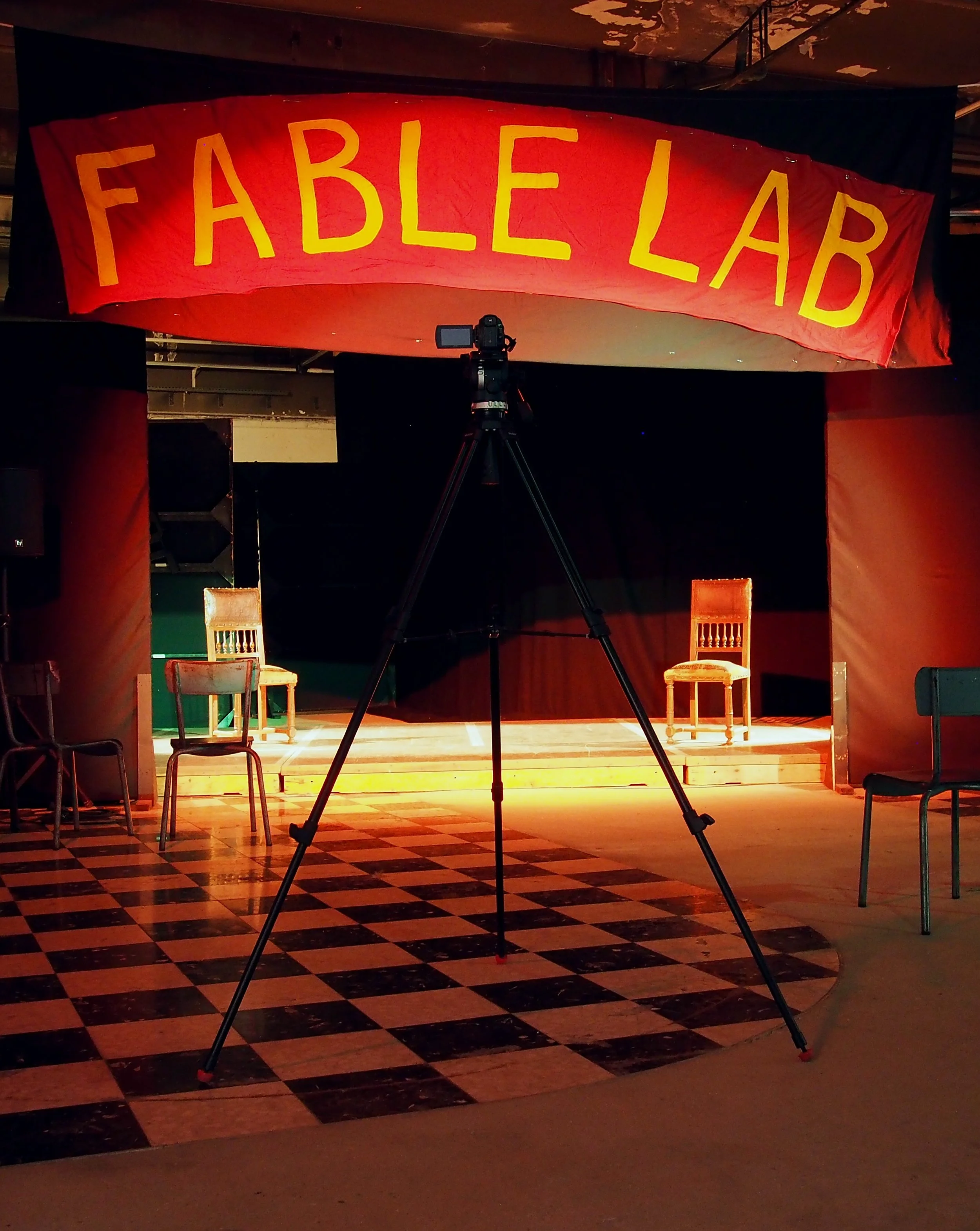 Scène de théâtre avec une scène éclairée et quatre chaises en bois, une caméra sur trépied est placée devant, une bannière rouge avec le mot 'FABLELAB' en jaune est suspendue en haut.