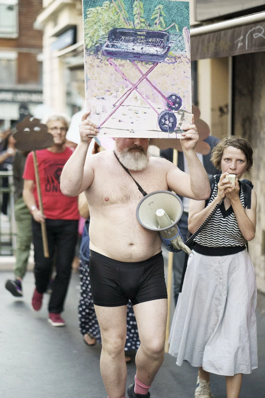 Un homme nu portant un mégaphone autour du cou et un tableau avec un piano accroché au-dessus de sa tête, lors d'une manifestation ou d'une marche, entouré par d'autres personnes.