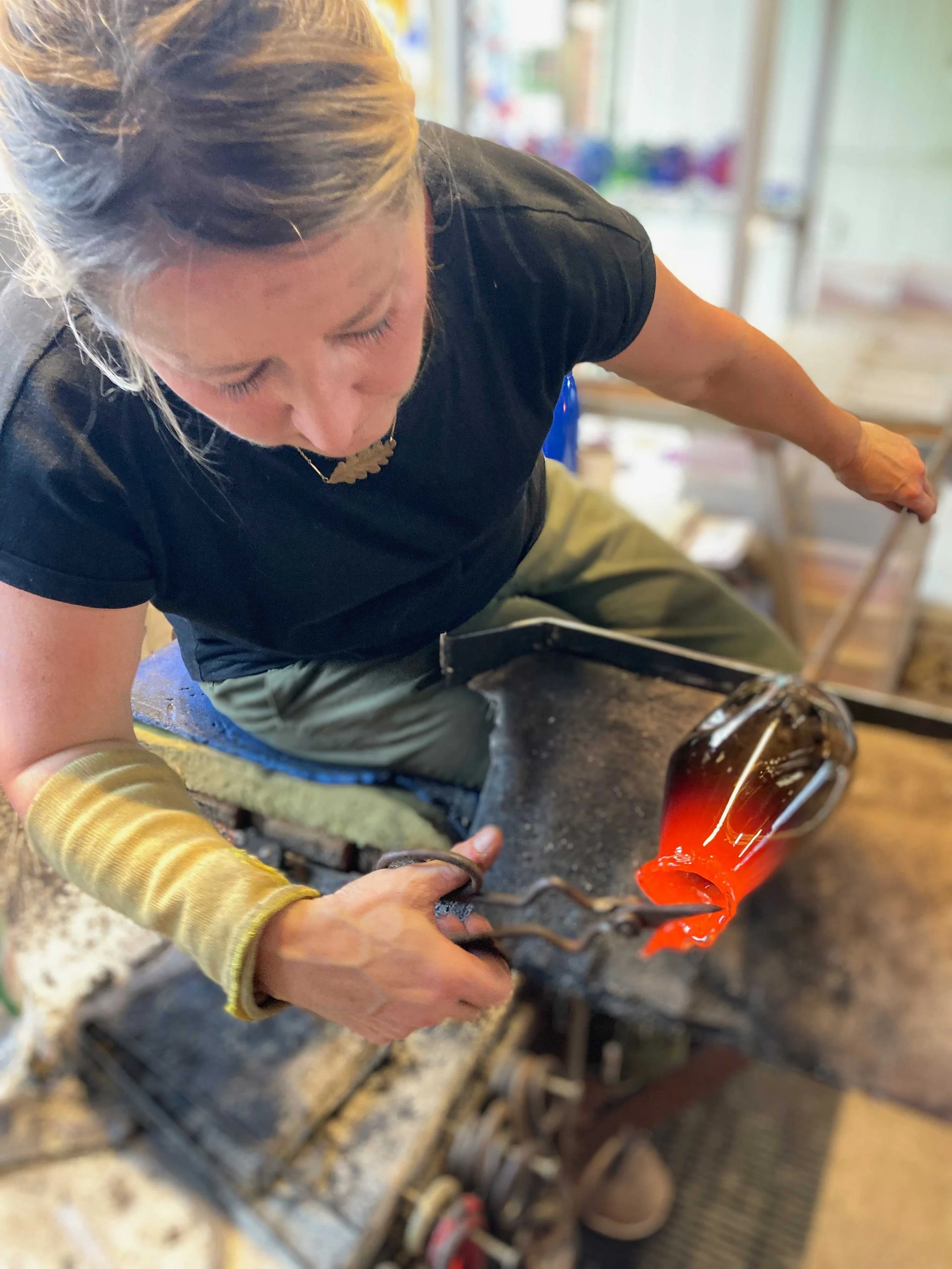 A woman pouring molten glass from a furnace, using tongs to handle the hot glass, with her hair tied back and wearing gloves for protection.