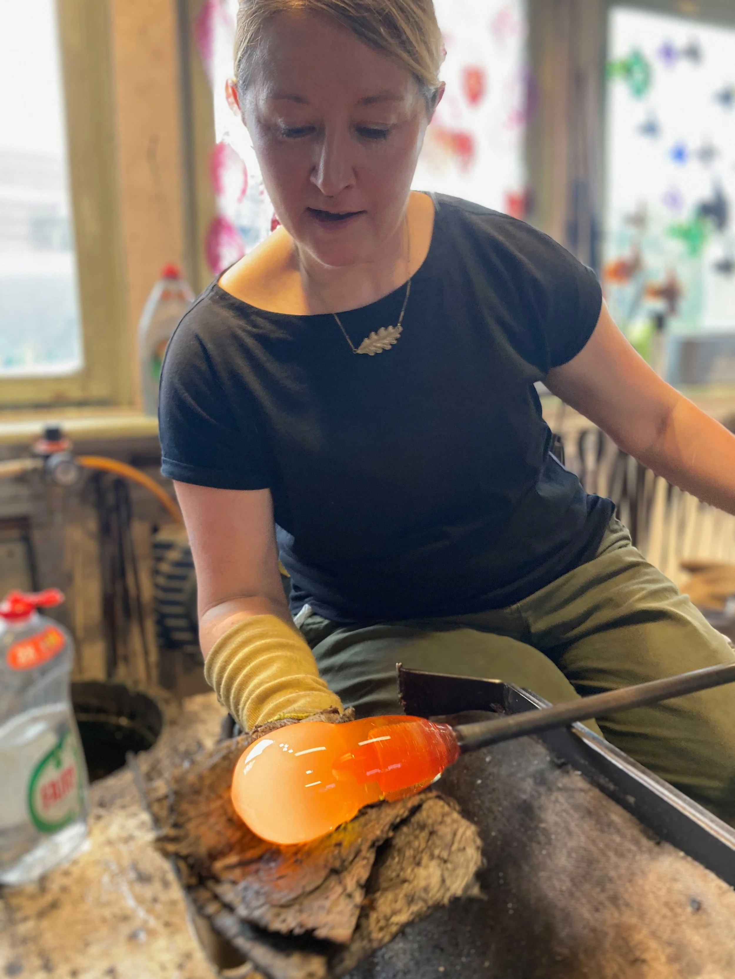A woman working with molten glass, shaping it on a workbench in a glassblowing studio.
