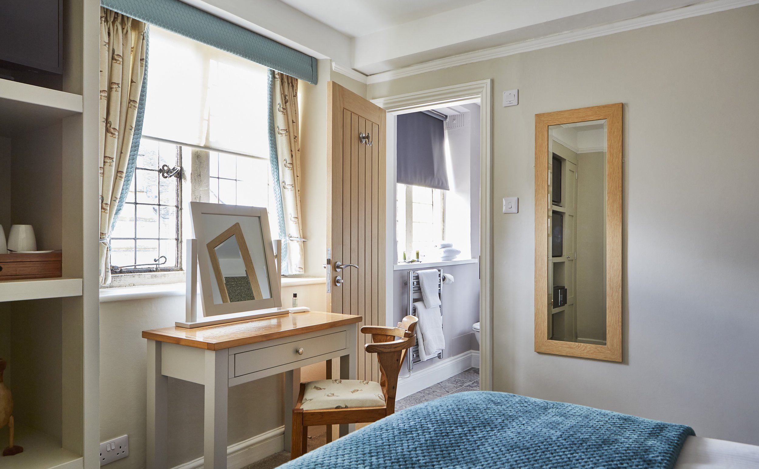Bedroom with a wooden vanity table and chair, wall-mounted mirror, window with patterned curtains, open door leading to bathroom, and blue bedding.