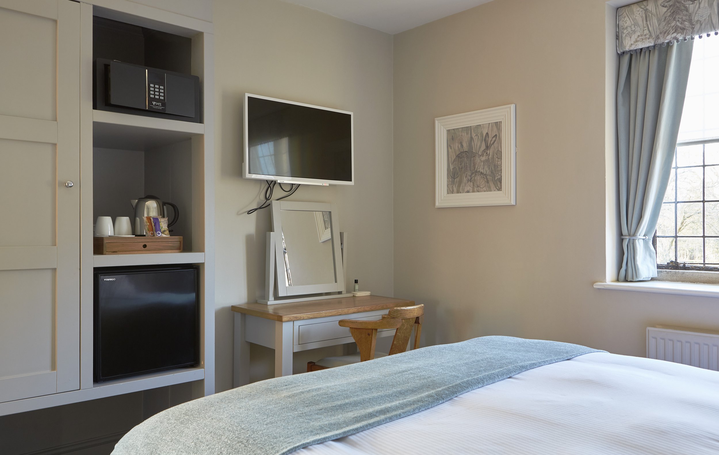 Hotel room interior with a bed, wall-mounted TV, desk, chair, framed artwork, and a built-in cabinet containing a mini fridge, safe, kettle, and cups. Window with curtains partially open.