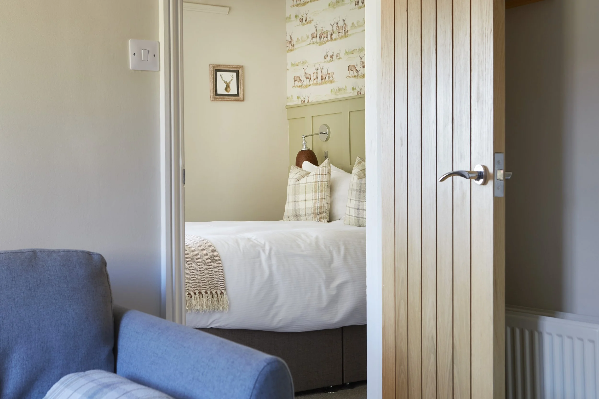 Cozy bedroom with open wooden door, white bedding, plaid pillows, and a blue armchair.