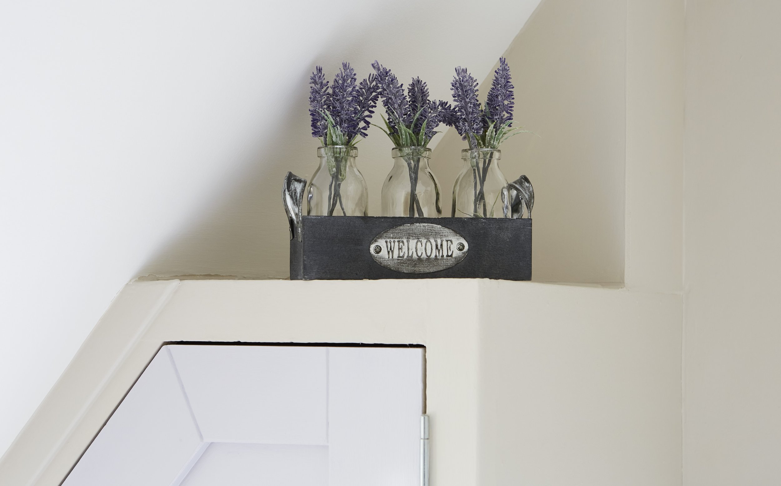 A decorative wooden box labeled 'WELCOME' containing three glass bottles with purple lavender stems, placed on a ledge against a white wall.