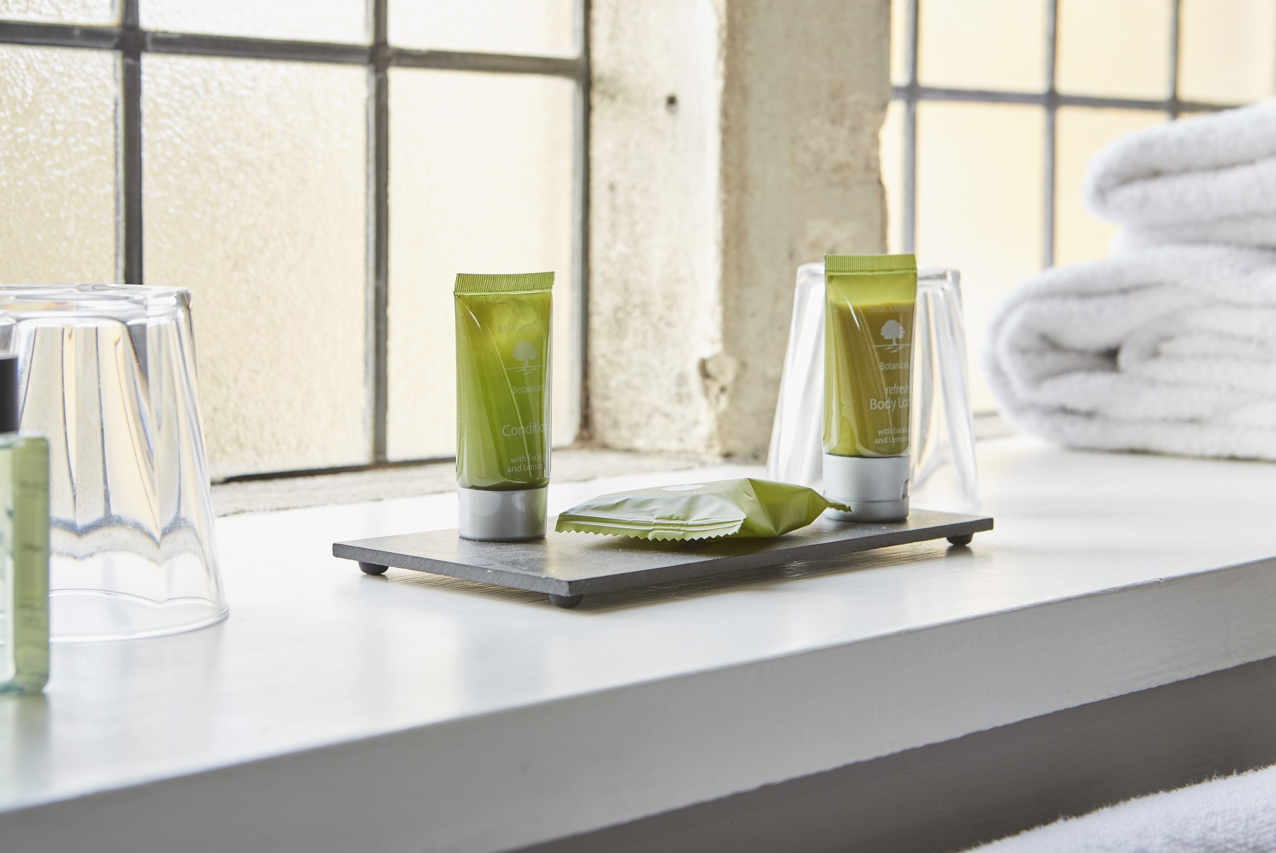 Bathroom countertop with travel-size toiletries like conditioner, body lotion, wrapped soap, clear plastic cups, and white towels near a textured window.