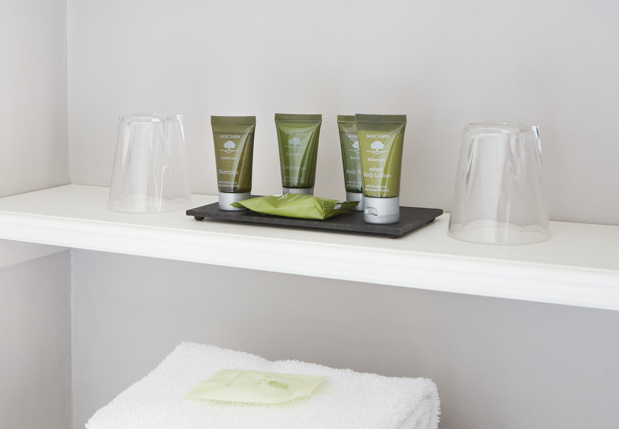 Hotel bathroom amenities on a shelf, including small bottles of shampoo, conditioner, body wash, and body lotion, with two plastic cups, a bar of soap, and a stack of towels underneath.