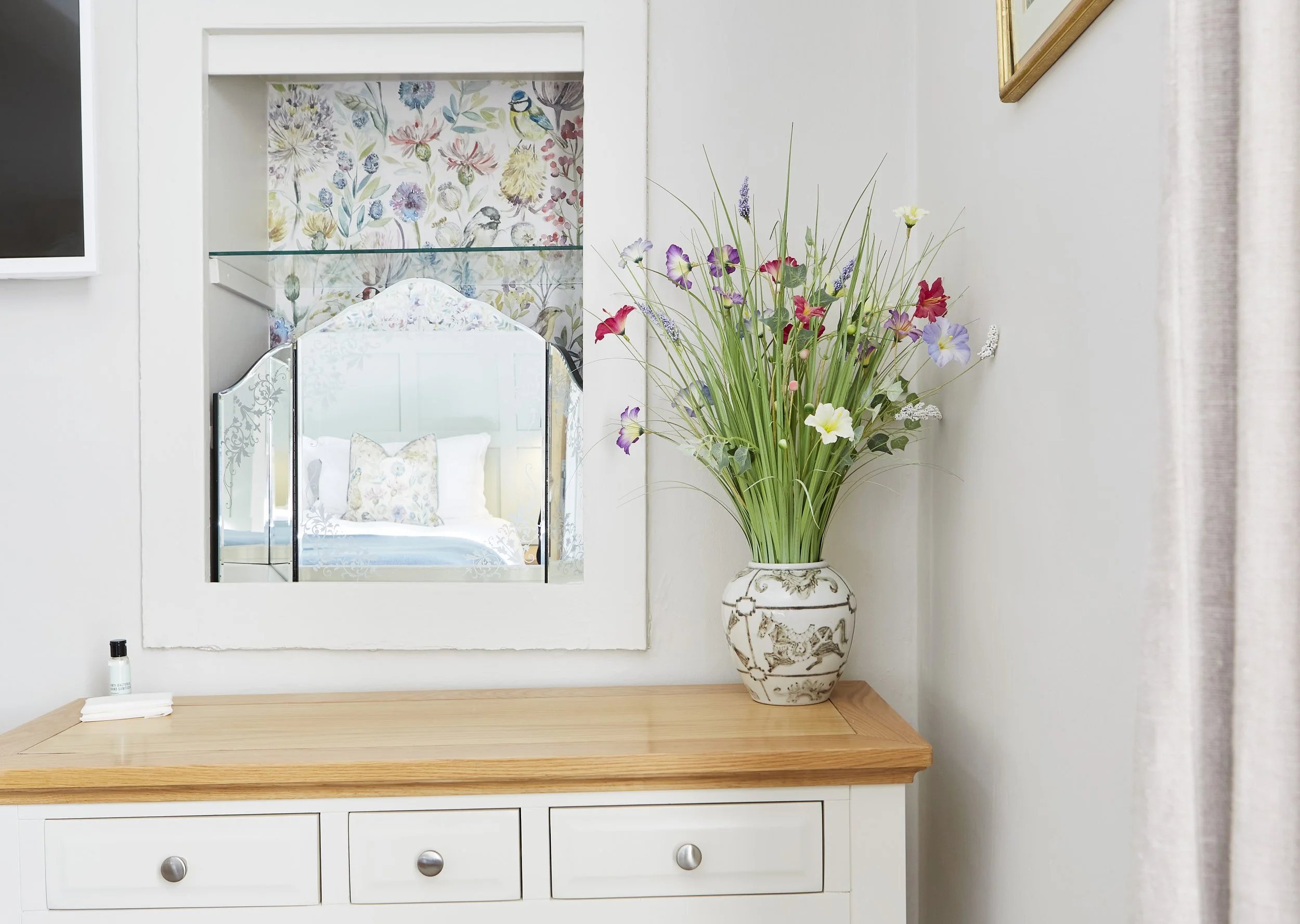 Room interior with a white dresser, decorative vase with flowers, and mirrored alcove reflecting floral wallpaper and part of a bed.