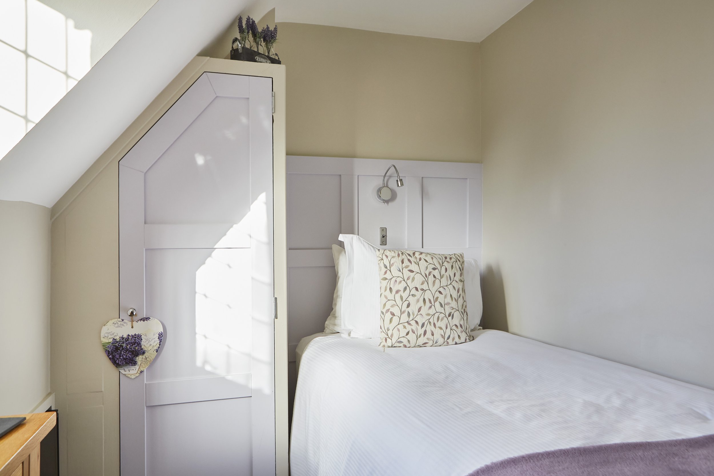 Cozy attic bedroom with a single bed, white linens, decorative floral pillow, angled ceiling, small closet, lavender decor item, and heart-shaped wall hanging.