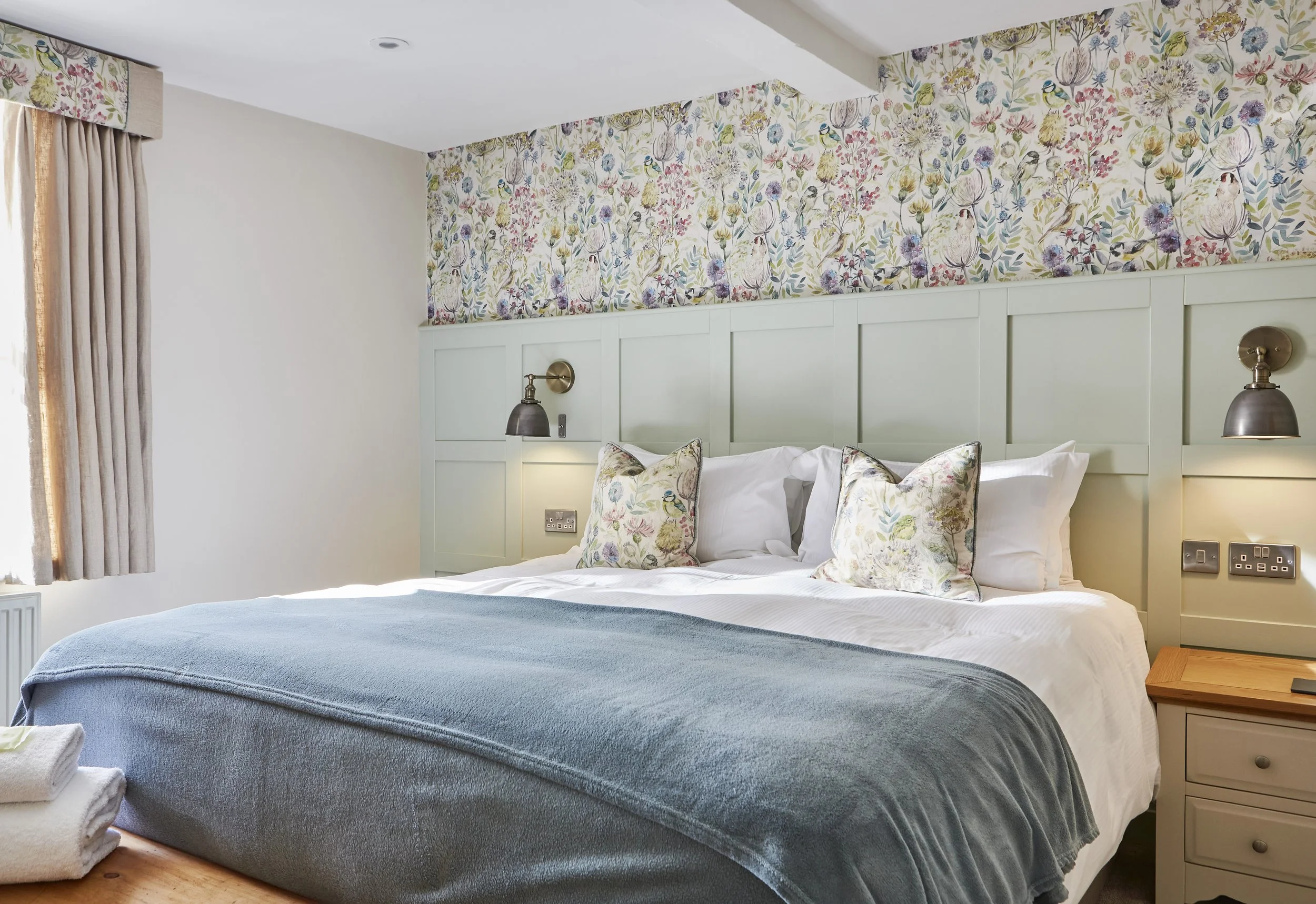 Cozy bedroom with a floral accent wall, king-sized bed with a blue blanket and floral pillows, wall-mounted lamps, and a curtain-covered window.