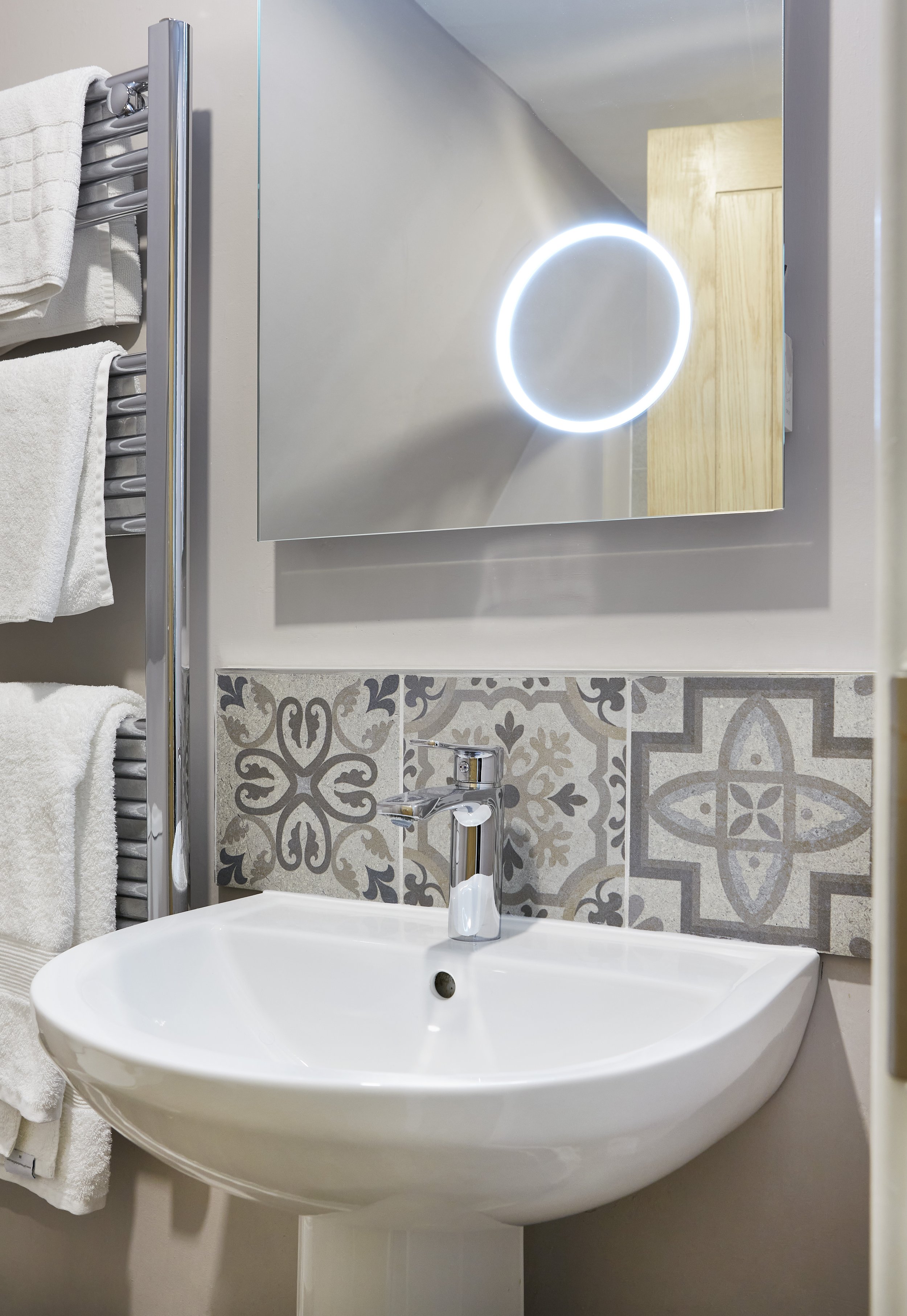 Modern bathroom with a white sink, decorative tiles, a wall-mounted mirror with LED light, and a towel rack with white towels.