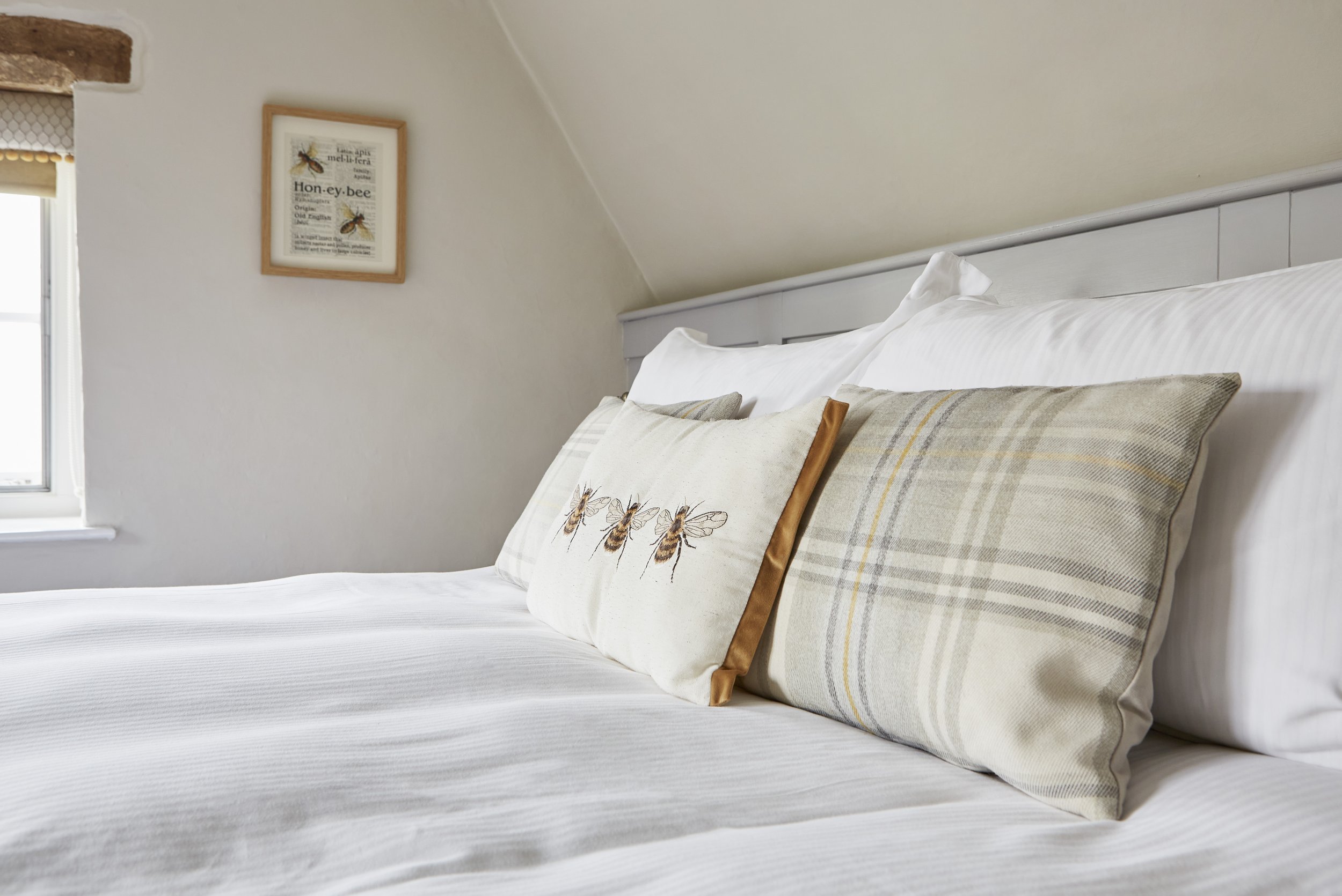 Cozy bedroom with a neatly made bed featuring bee-themed and plaid cushions, a light-colored headboard, and honey bee artwork on the wall.
