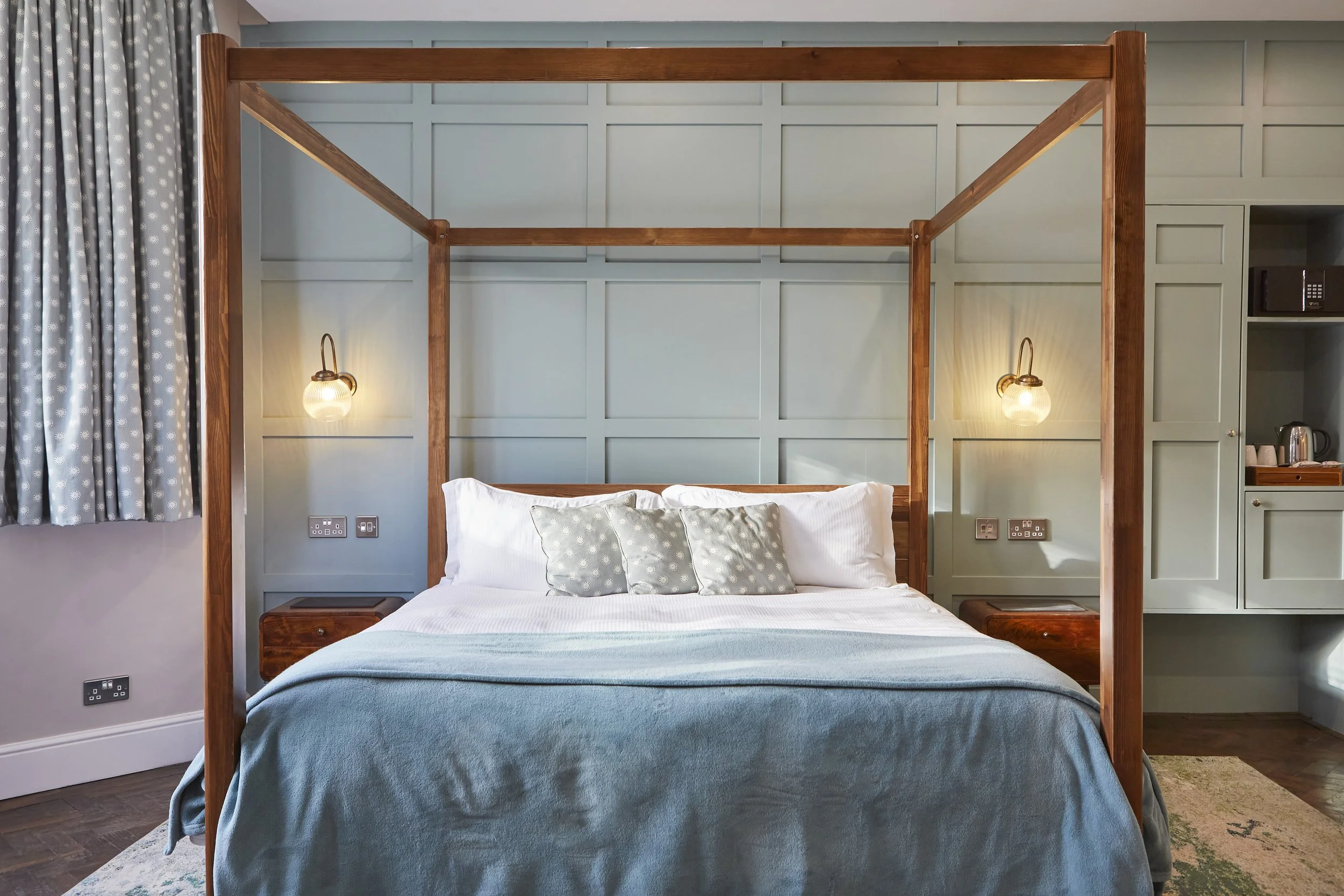 Elegant bedroom with a four-poster bed, light blue bedding, patterned pillows, and wall sconces. A paneled wall, gray patterned curtains, and bedside tables complete the decor.