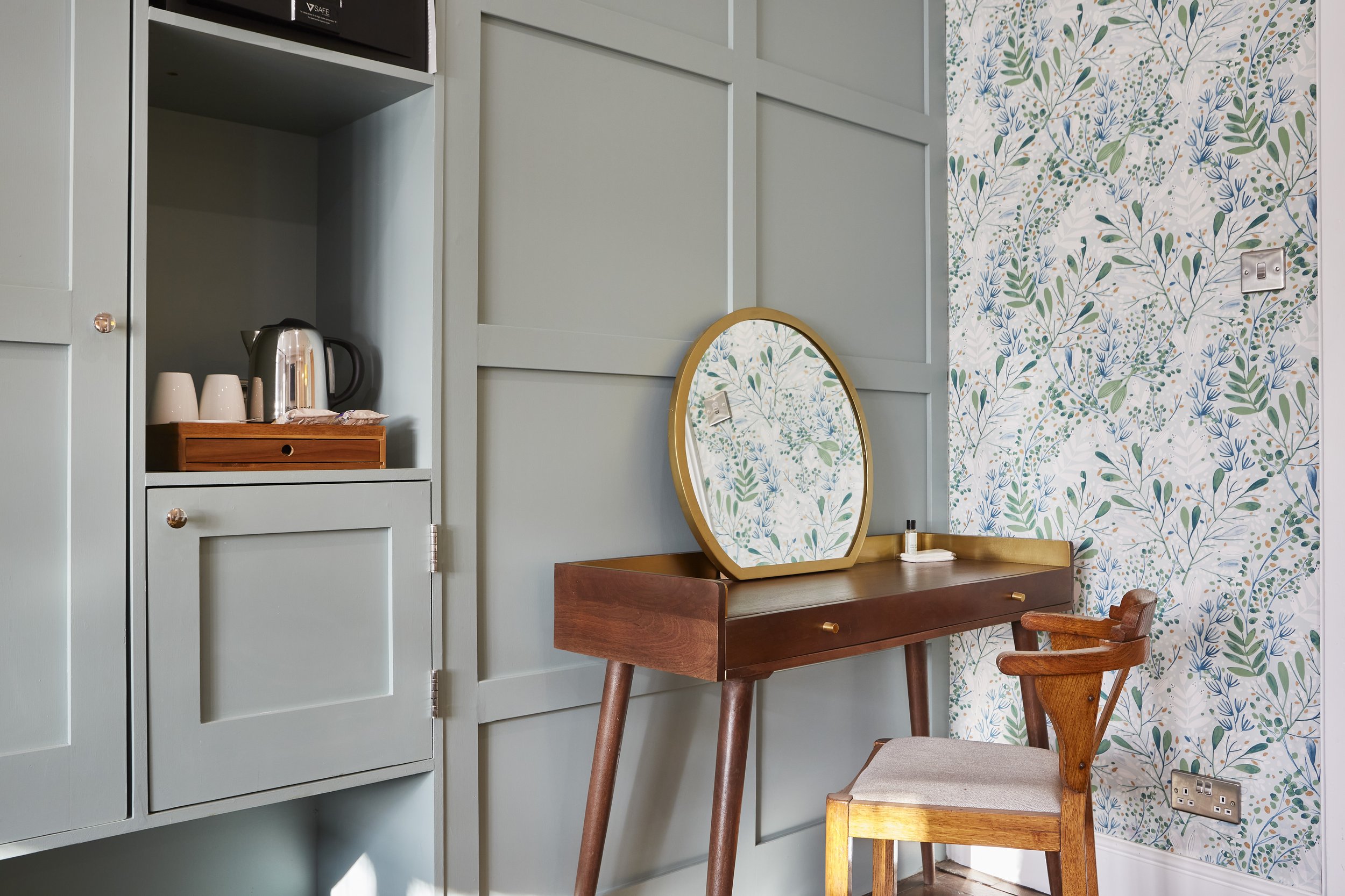 Room interior with a wooden desk next to a green paneled wall. The desk holds a circular mirror with floral wallpaper reflecting in it. There's a vintage wooden chair in front of the desk. A built-in cabinet with a kettle and cups is visible on the l