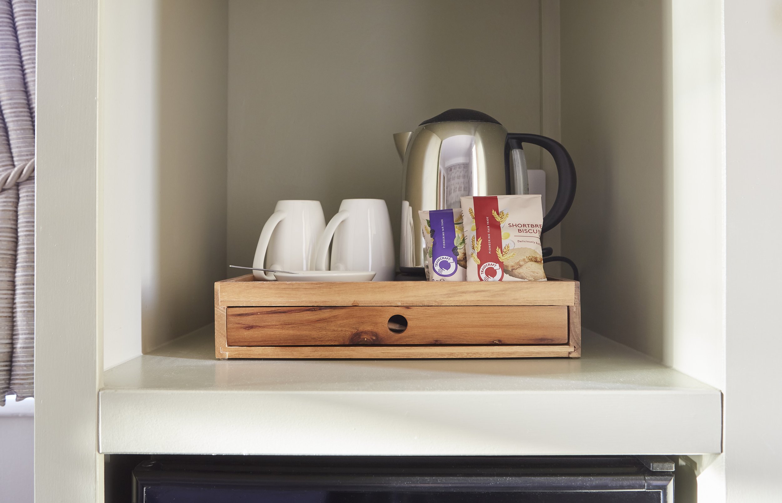 Hotel room tray with tea and coffee amenities, including white mugs, electric kettle, spoon, and shortbread biscuits on a wooden tray.