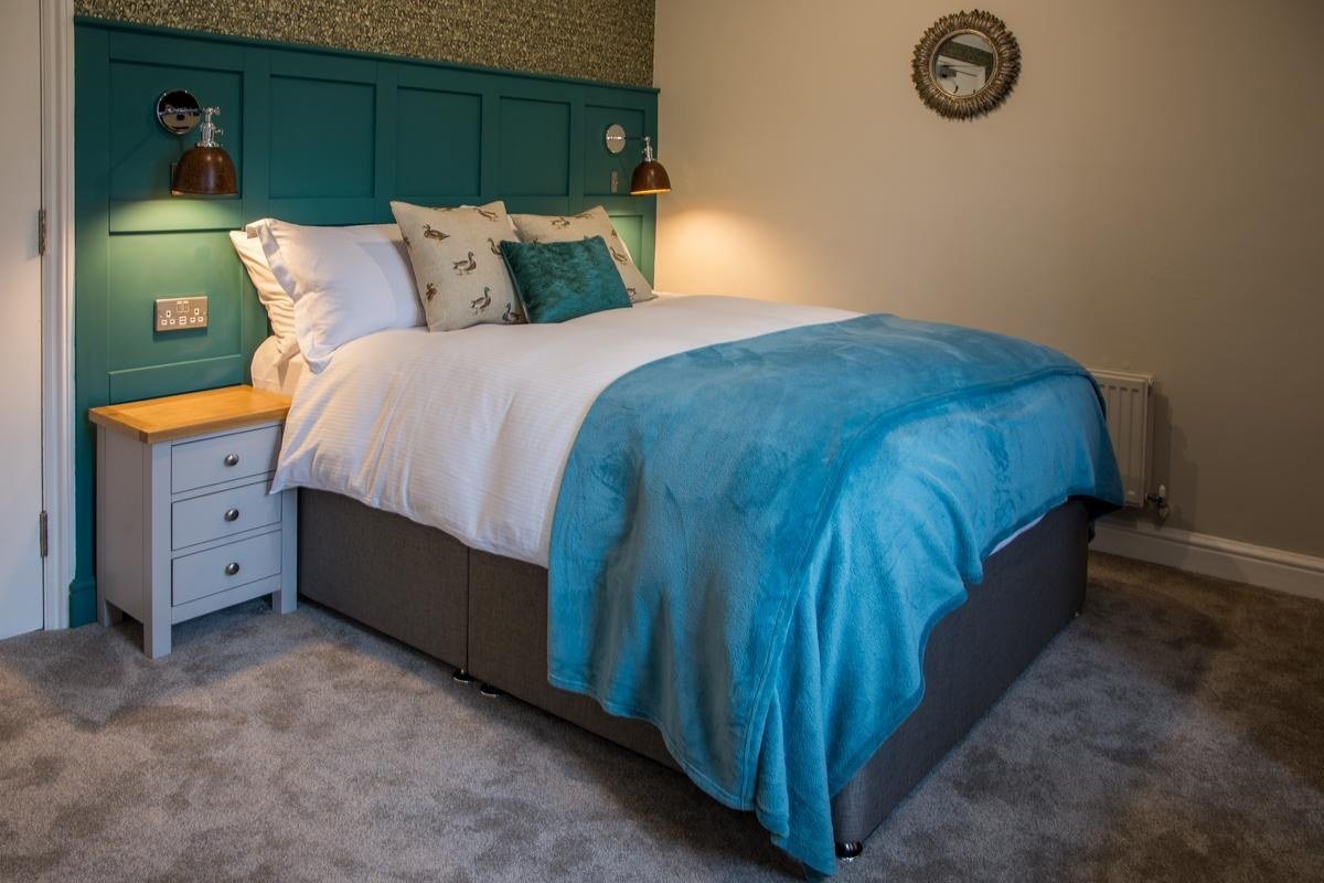 Cozy bedroom with a large bed featuring white linens, teal throw blankets, and decorative pillows. The headboard is teal with two wall-mounted lamps. A small bedside table with drawers is on the left. A circular mirror hangs on the beige wall.