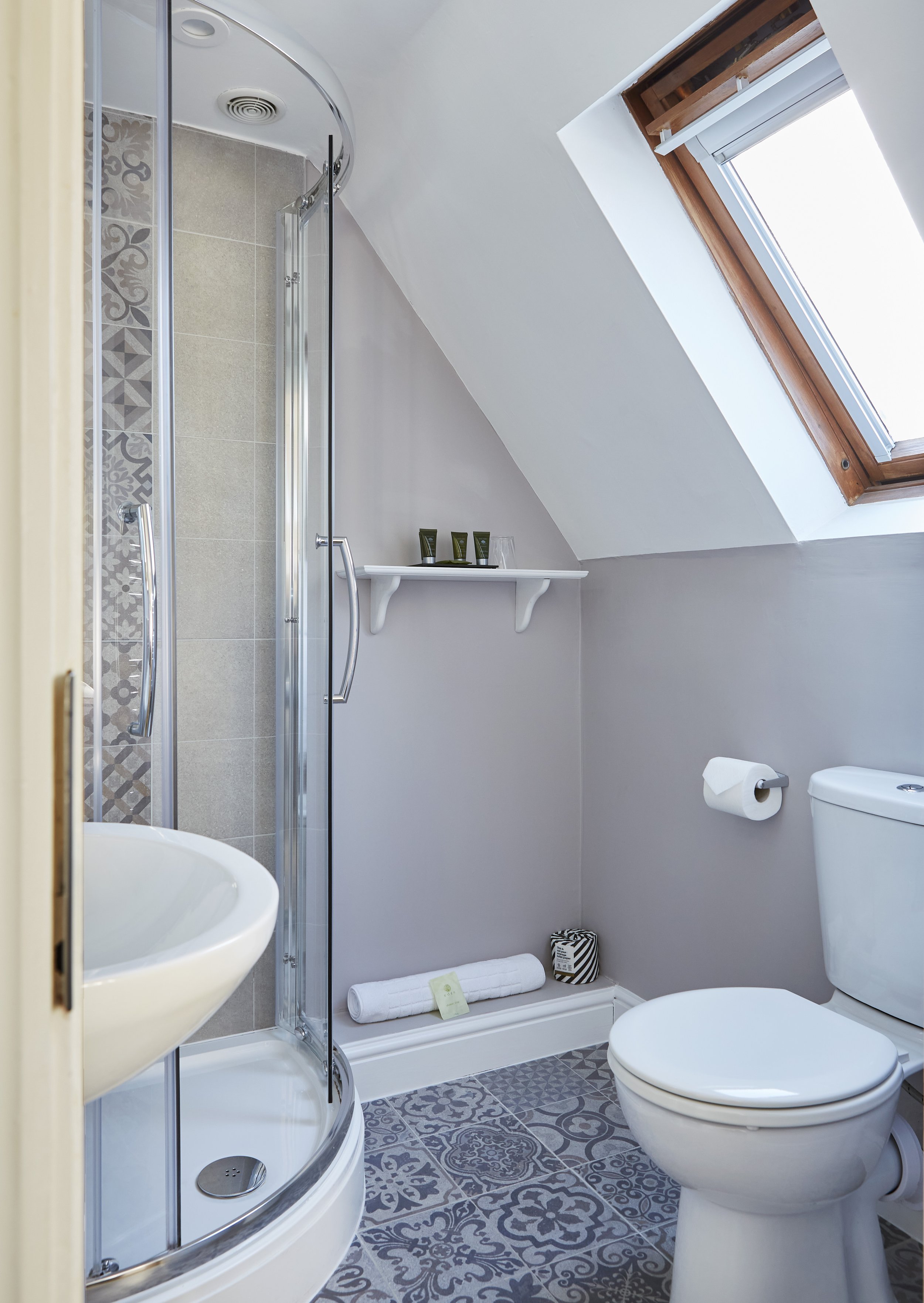 Small modern bathroom with circular shower, white toilet, pedestal sink, gray walls, patterned tile floor, shelf with toiletries, and skylight window.