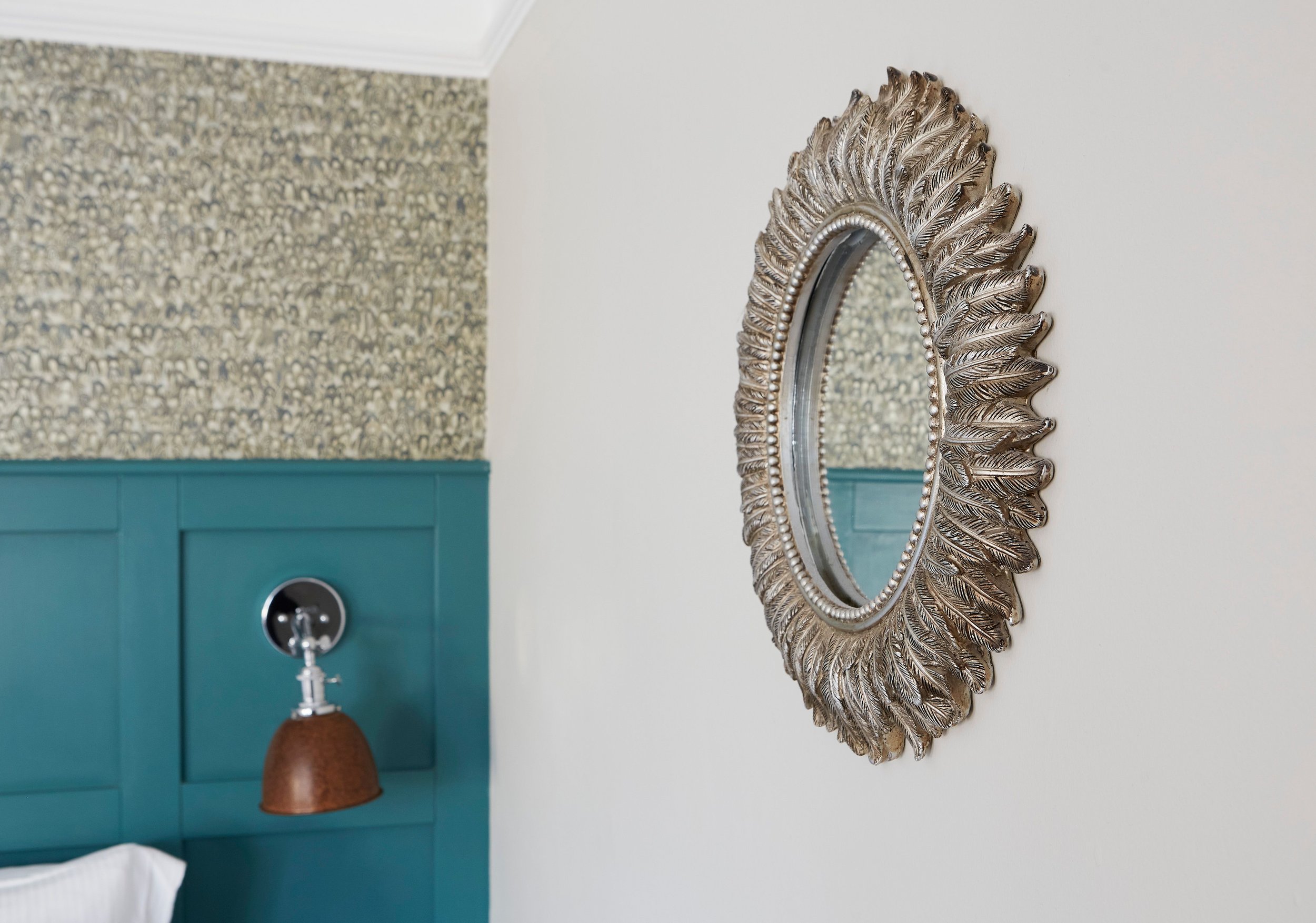 Decorative mirror with ornate frame on wall next to blue paneling and wall sconce.