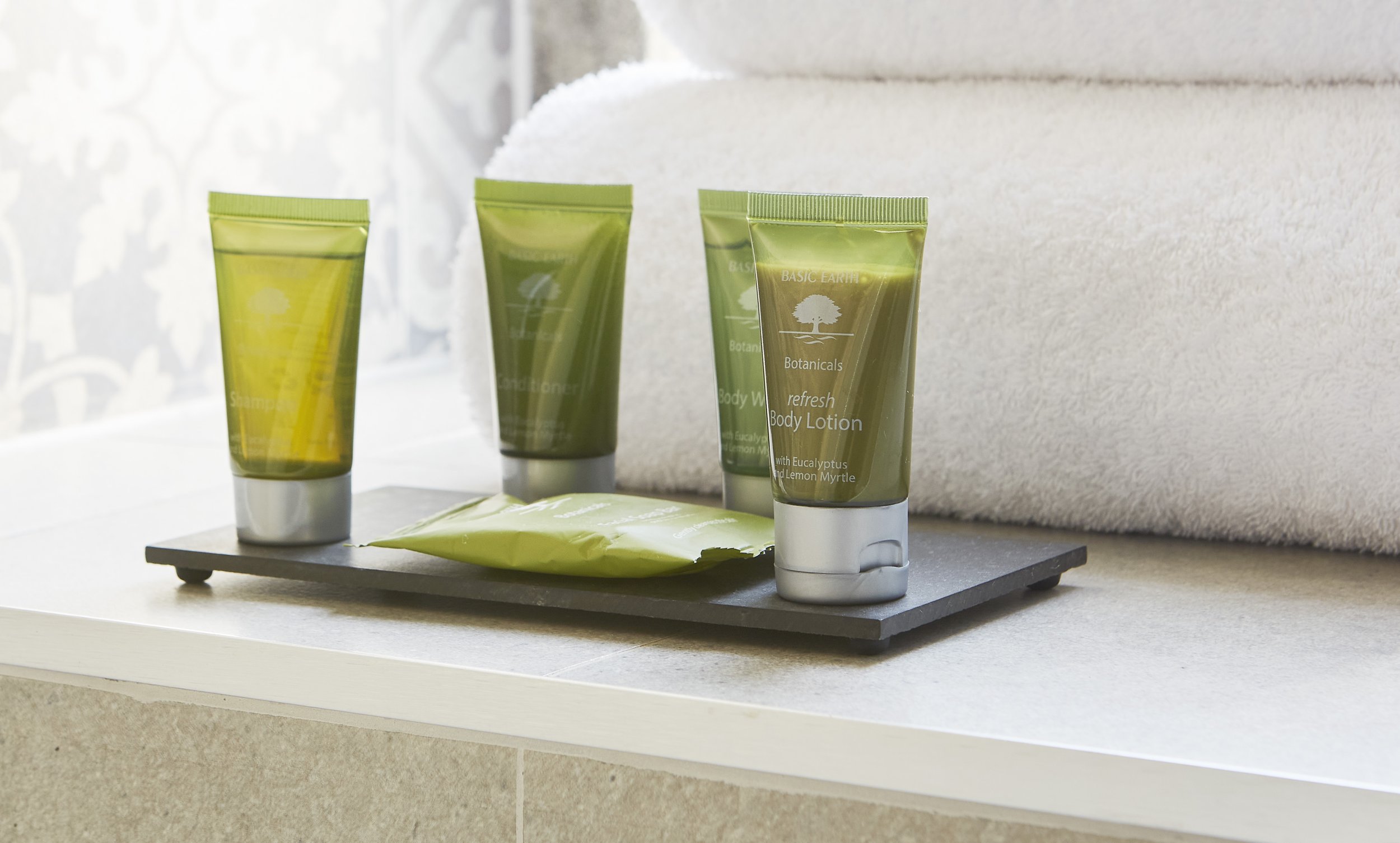 Hotel toiletries including shampoo, conditioner, body wash, and body lotion in green tubes, displayed on a tray next to folded white towels.