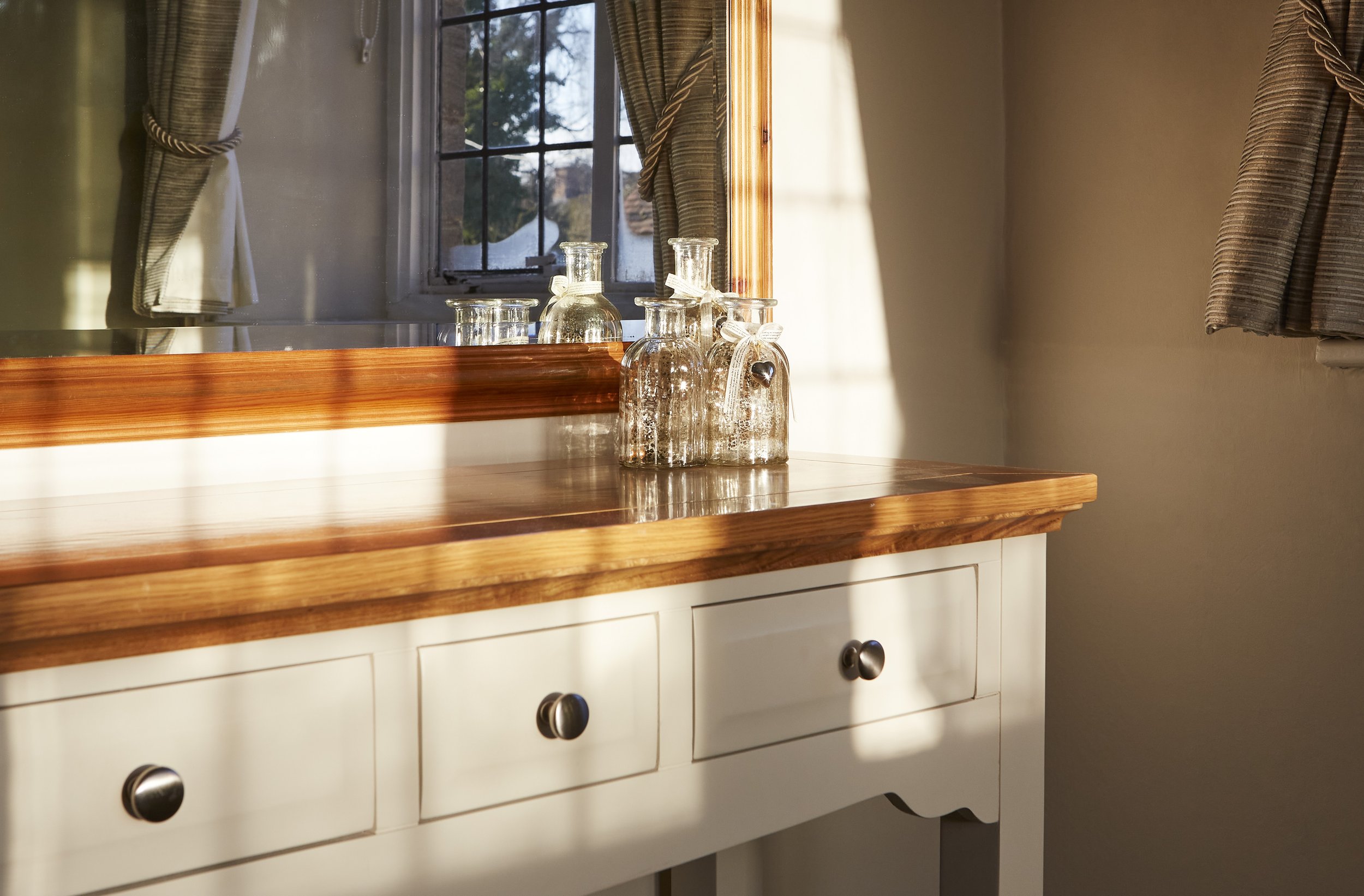 Wooden dresser with glass bottles, mirror, and curtains in a sunlit room.