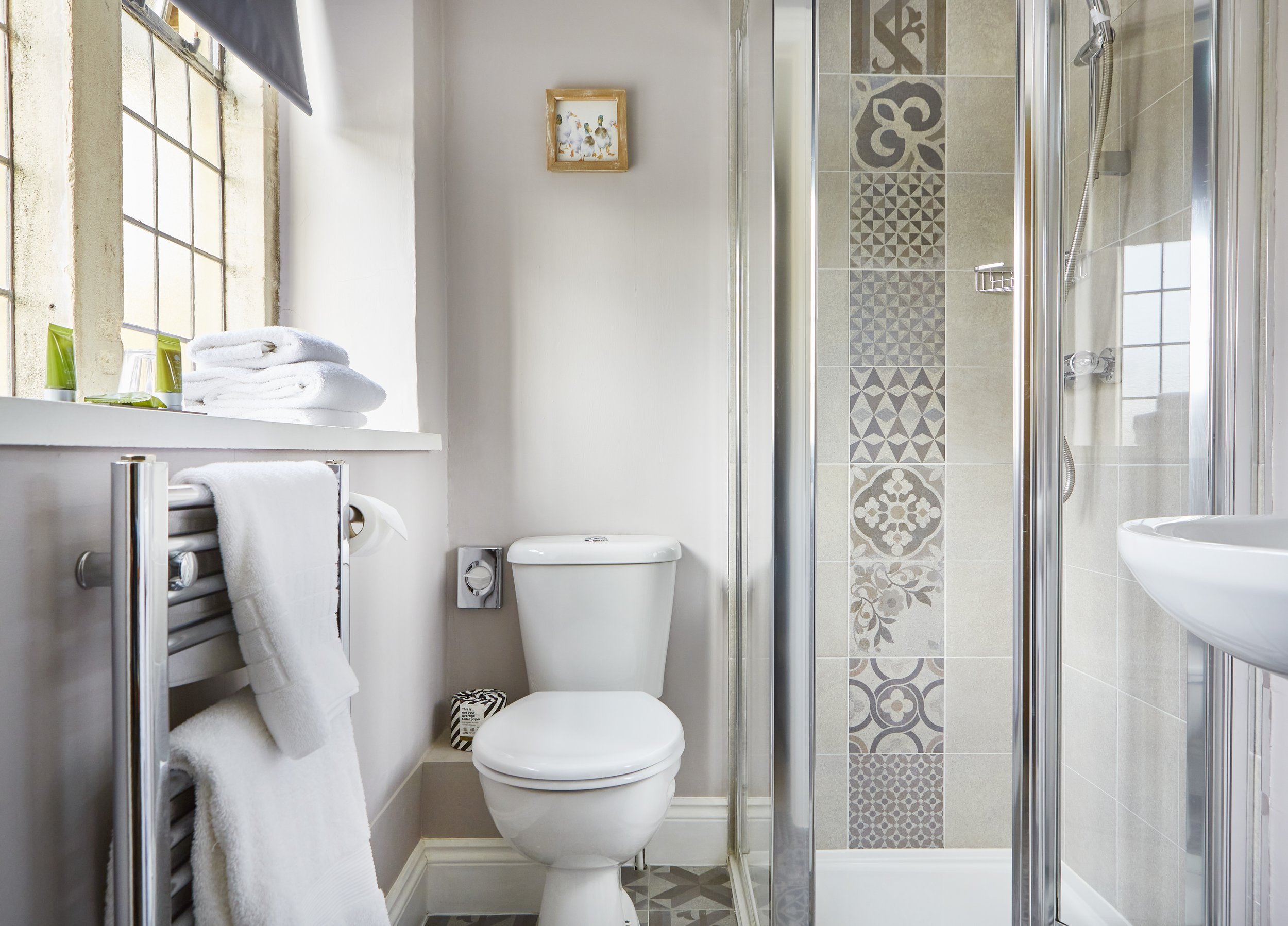 Modern bathroom with patterned tile shower, white toilet, radiator with towels, and small decorative items on the windowsill.