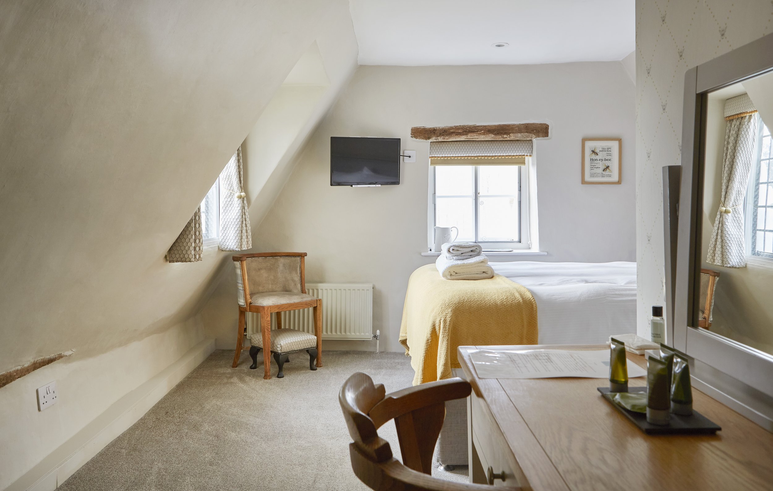 Cozy attic bedroom with slanted ceiling, bed with yellow blanket, stacked towels, wooden chair, wall-mounted TV, desk with toiletries, and a small window.