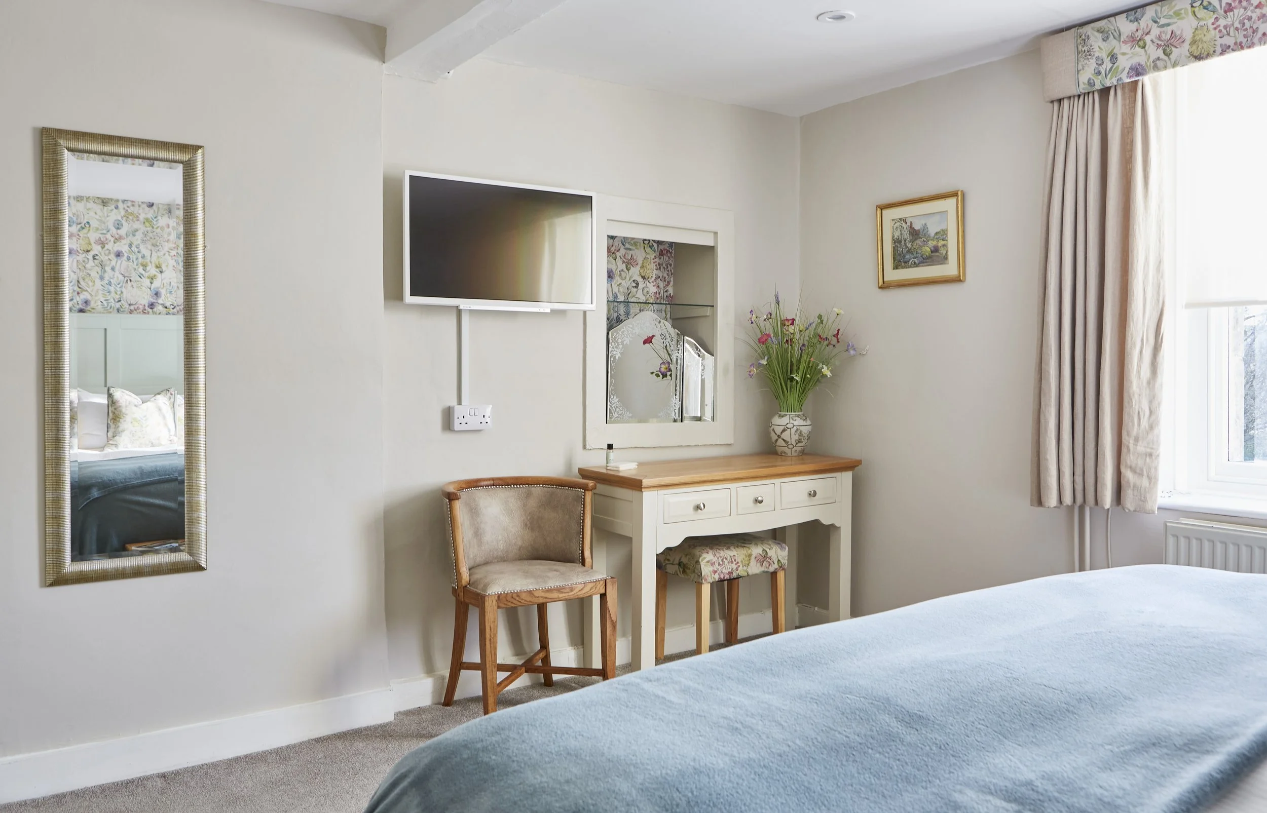 Bedroom interior with a wall-mounted TV, a wooden desk with floral arrangement, an upholstered chair, a decorative mirror, and a window with curtains.