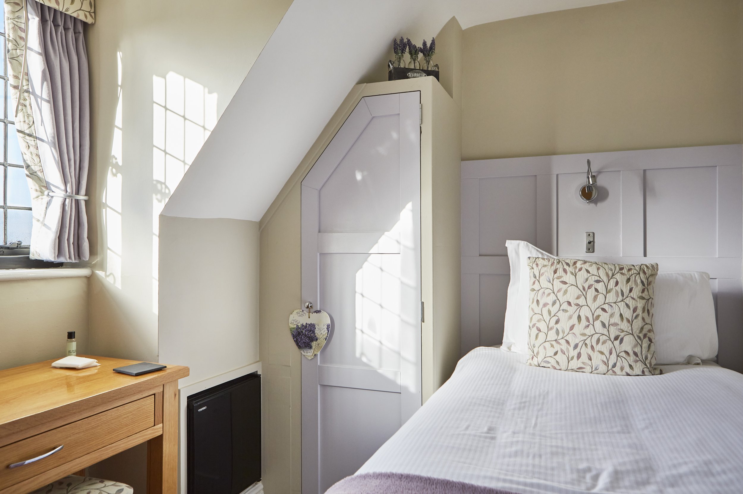 Cozy bedroom with a single bed, floral pillow, and a purple throw. A wooden desk with a smartphone and lotion sits by a window with draped curtains. A sloped ceiling and a small closet with a heart-shaped ornament are visible.