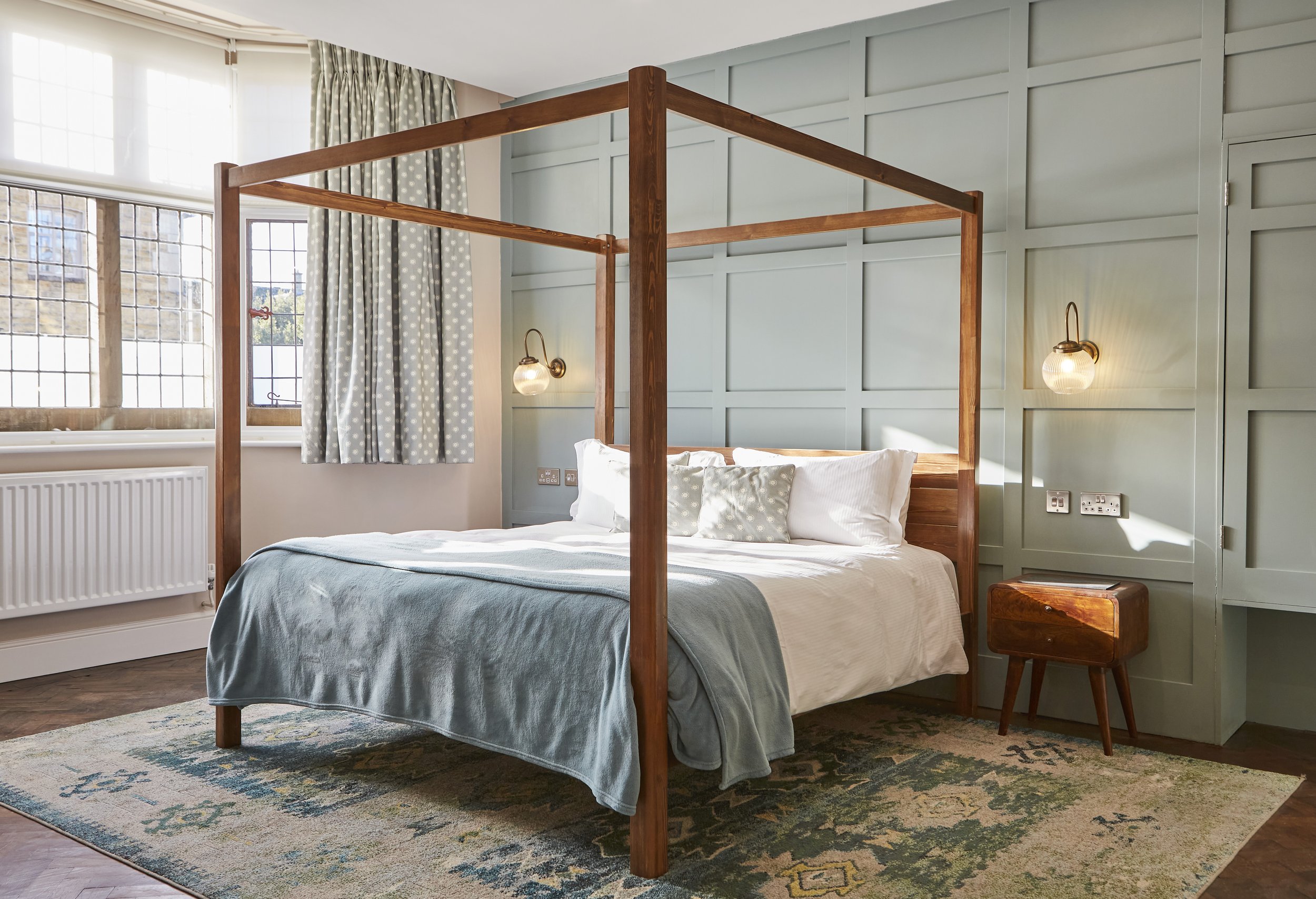 Bedroom with canopy bed, light blue blanket, patterned rug, paneled walls, bedside table, wall sconces, and large windows with curtains.