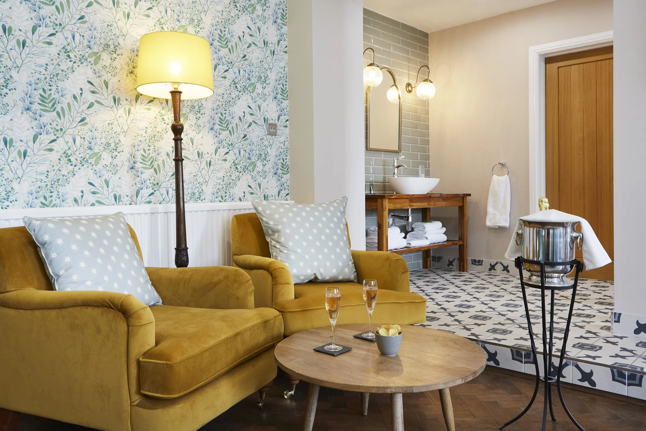 Cozy living area with mustard-yellow armchairs, a round wooden table, and a floor lamp. There's a floral patterned wallpaper, a small open bathroom area with a sink and towels, and a champagne bucket with a bottle on a stand. The space has a warm and