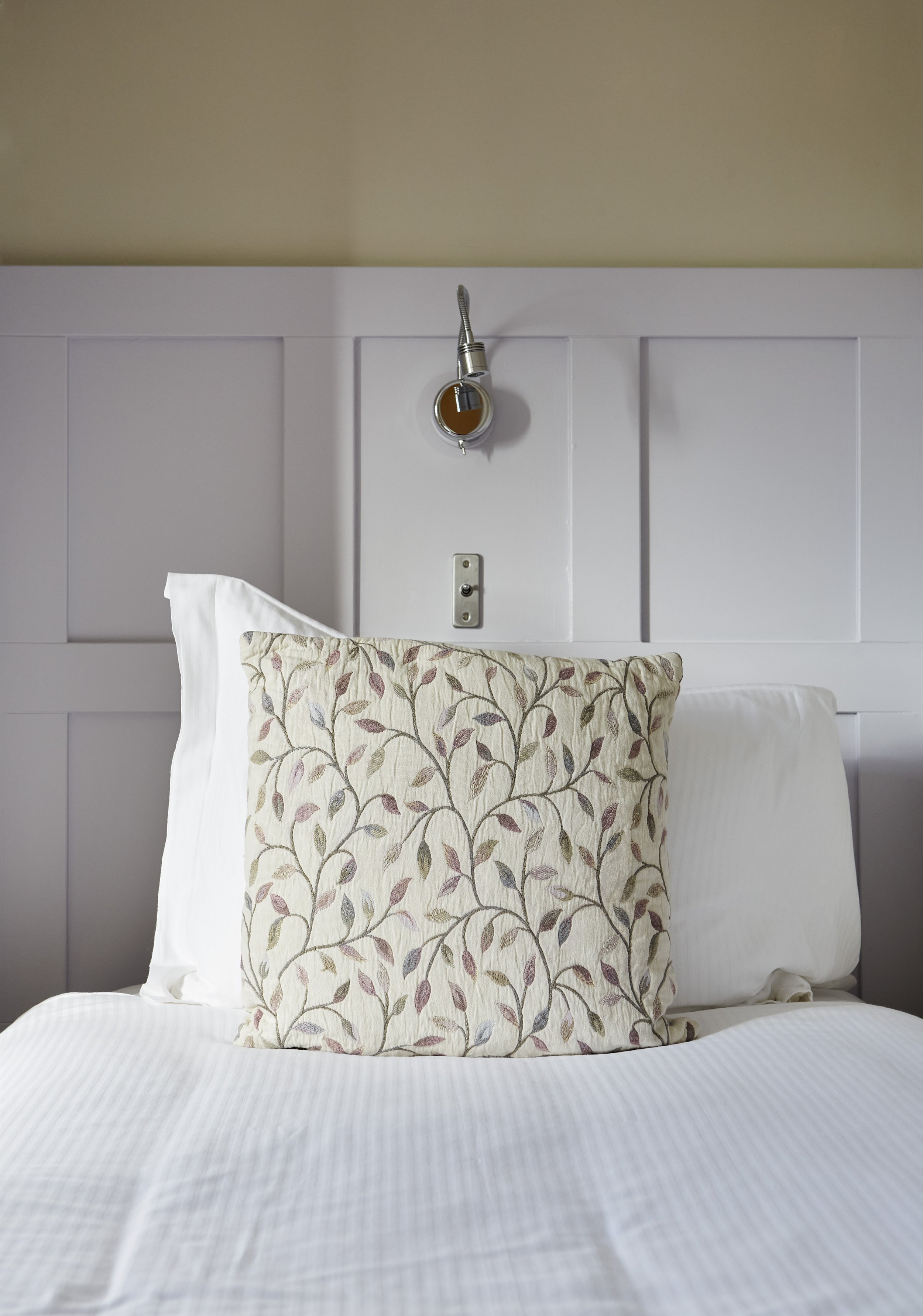 A bed with white pillows and a decorative pillow with a leaf pattern, set against a white paneled headboard with a wall-mounted reading lamp above.