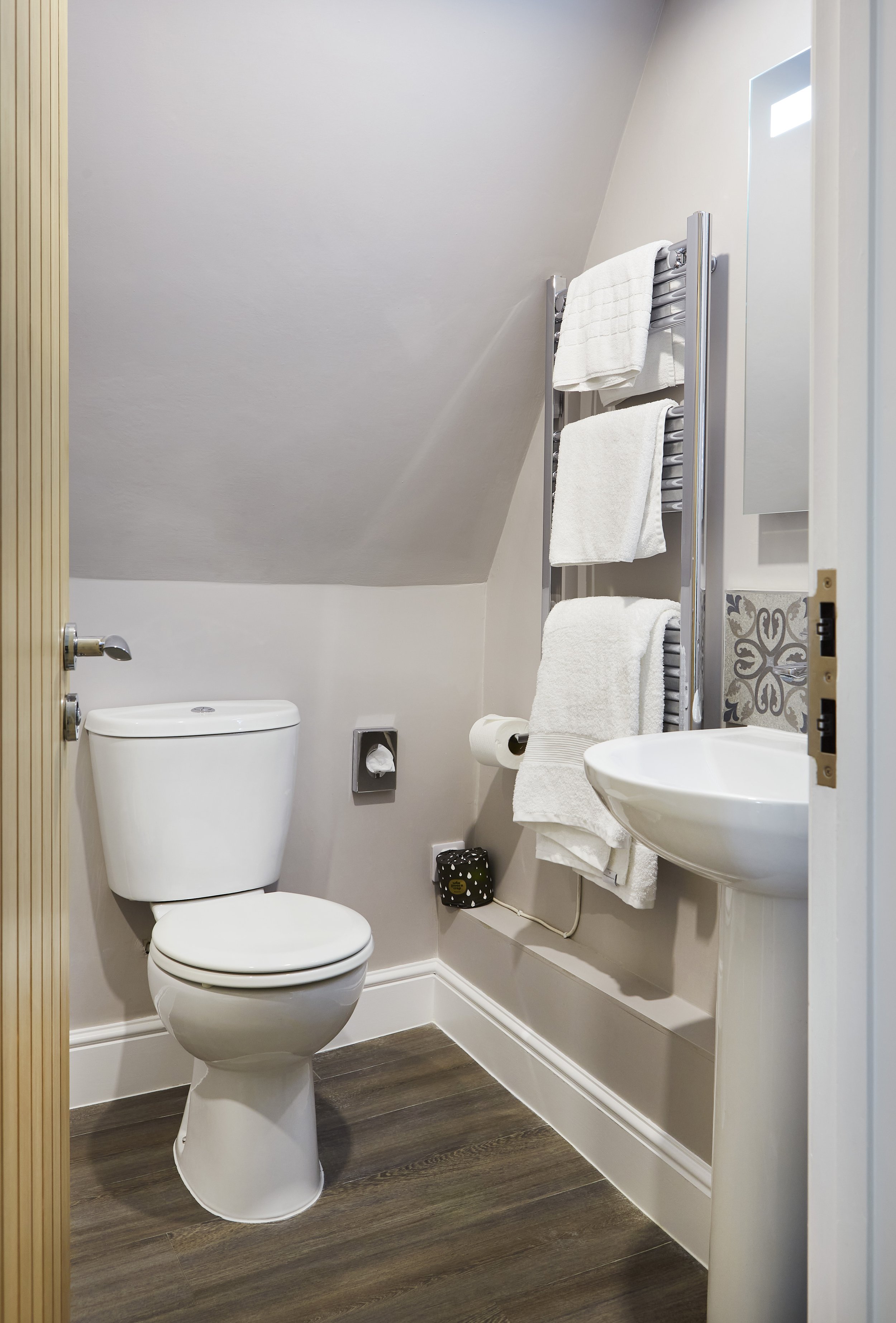 Modern small bathroom with a toilet, pedestal sink, and a wall-mounted towel rack holding white towels. The room has light-colored walls and wood-style flooring.