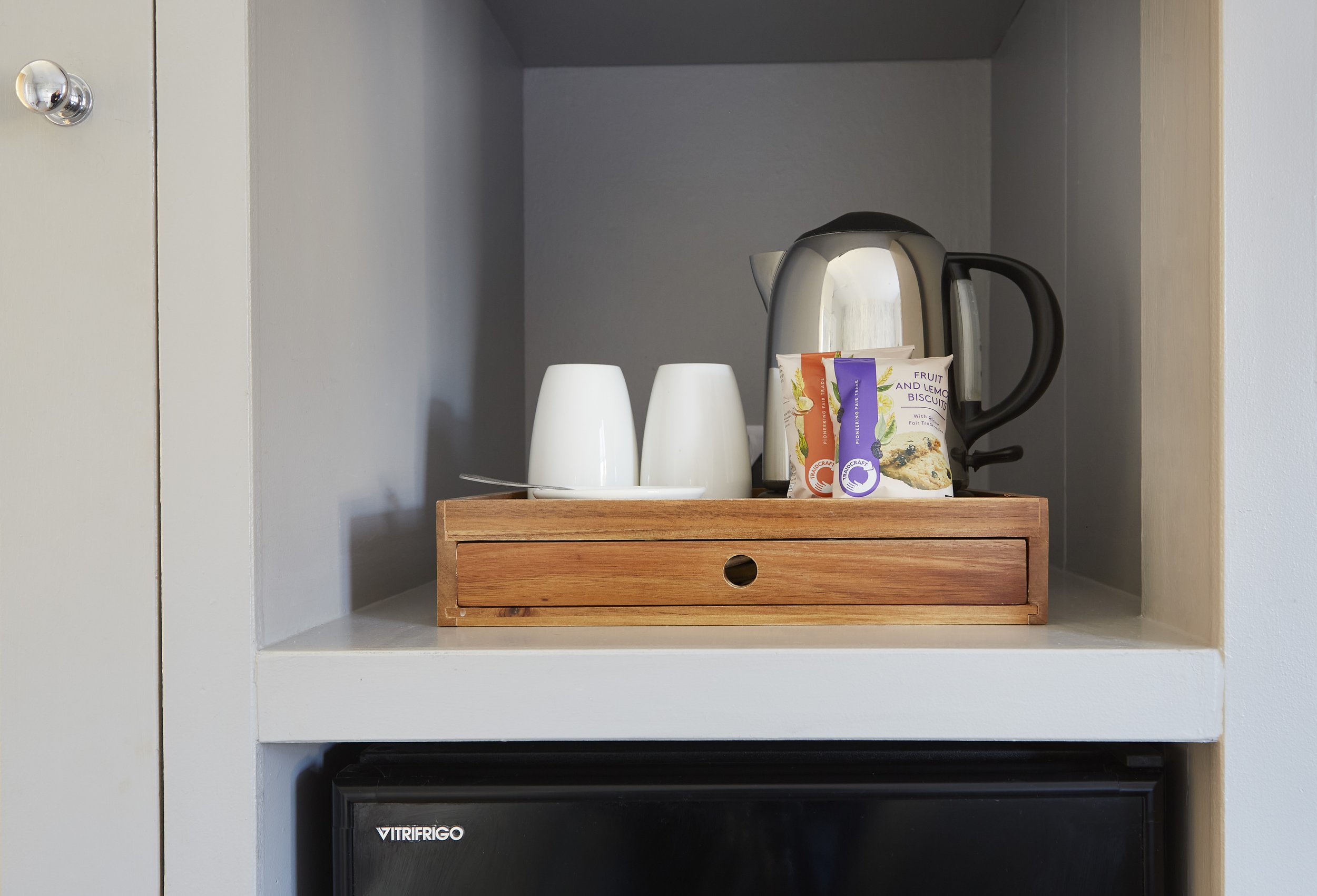 Small kitchenette setup with a kettle, two white cups, a snack pack, and a wooden drawer on top of a mini fridge in a compact shelf space.